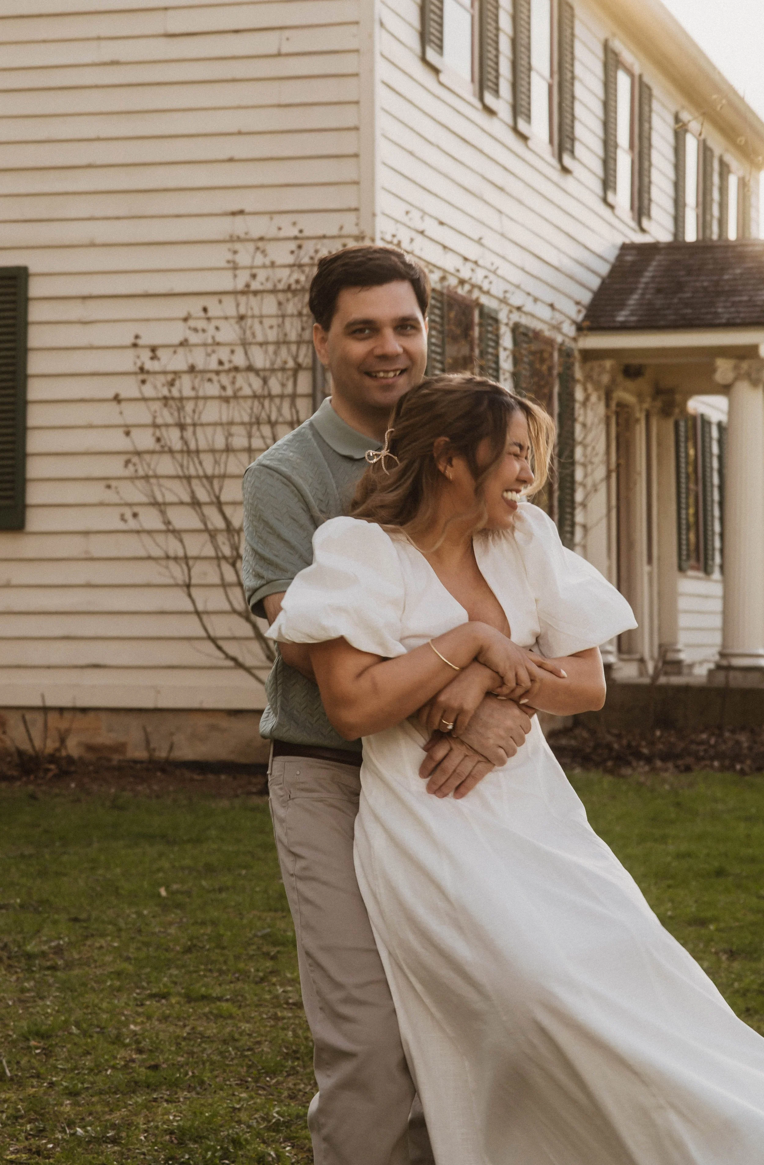 A man and a woman happily embracing outdoors in front of a white house with black shutters, during sunset. The woman is in a white dress and the man is in a light-colored shirt and pants.