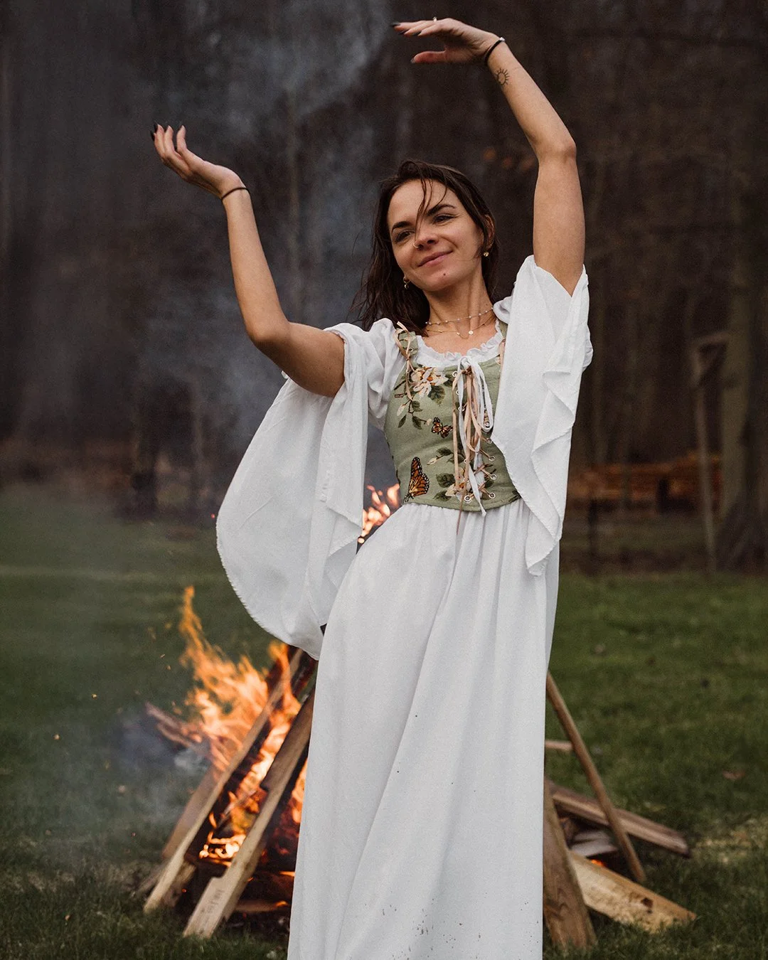 A woman stands outdoors near a bonfire, dancing with her arms raised above her head, smiling softly. She is wearing a white skirt, a green top with butterfly designs, and a white open shirt. The scene is set in a grassy area with trees in the backgro