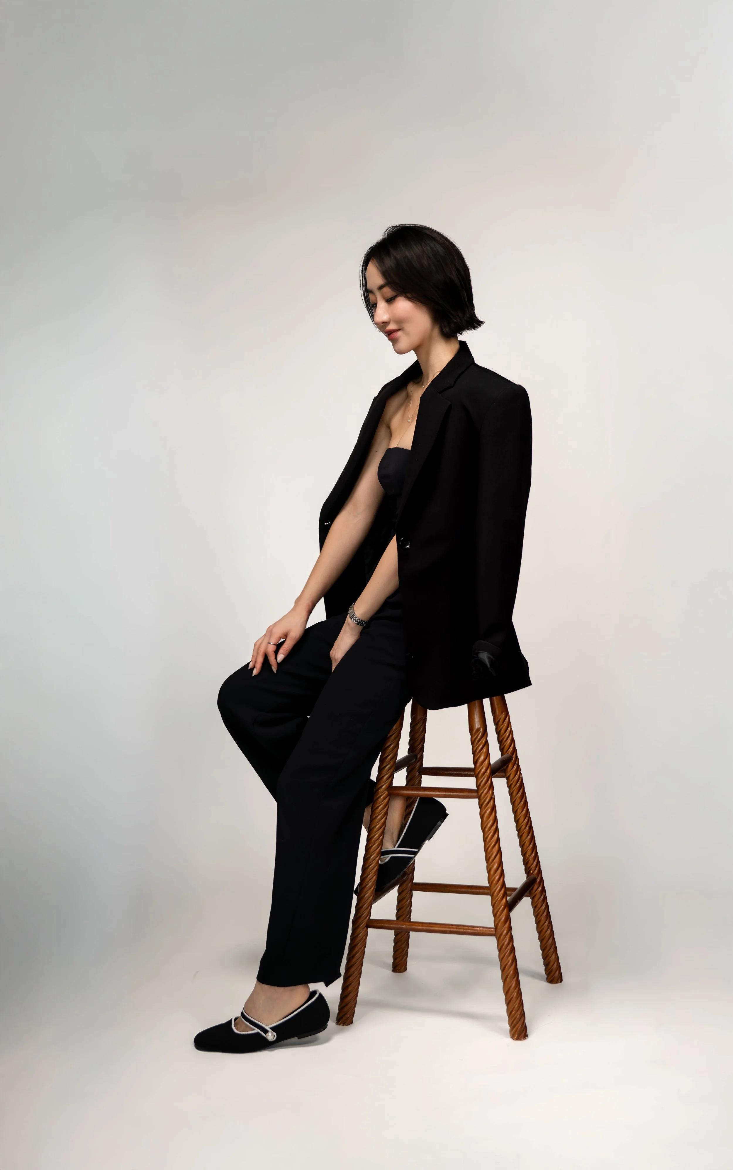 A woman sitting on a wooden stool draped in a black blazer, black top, and black pants, with black and white shoes, posing against a plain white background.
