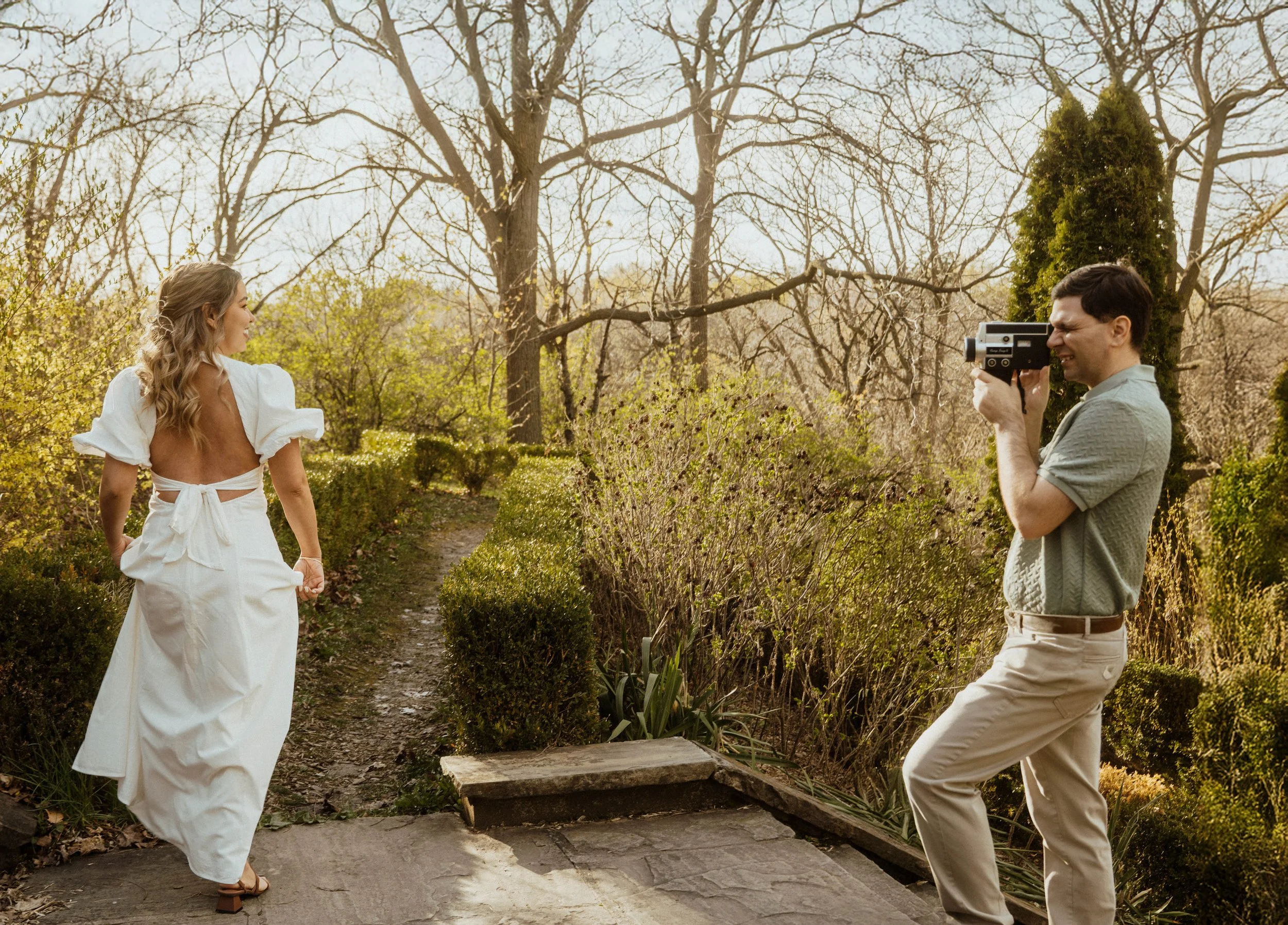 A woman in a white dress posing outdoors while a man takes her photograph with a vintage camera