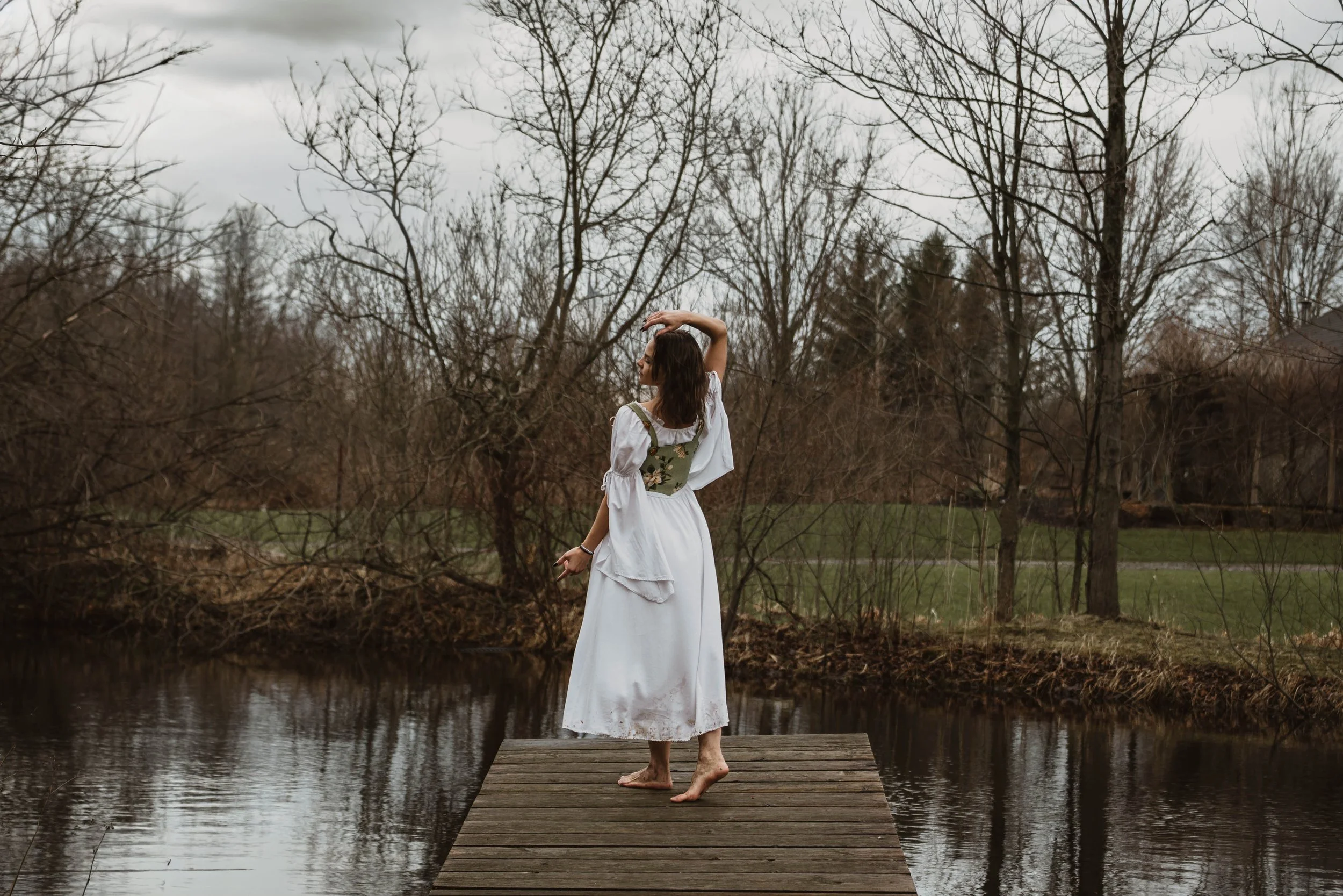 A woman in a white dress standing barefoot on a wooden dock by a body of water, with bare trees in the background under an overcast sky.