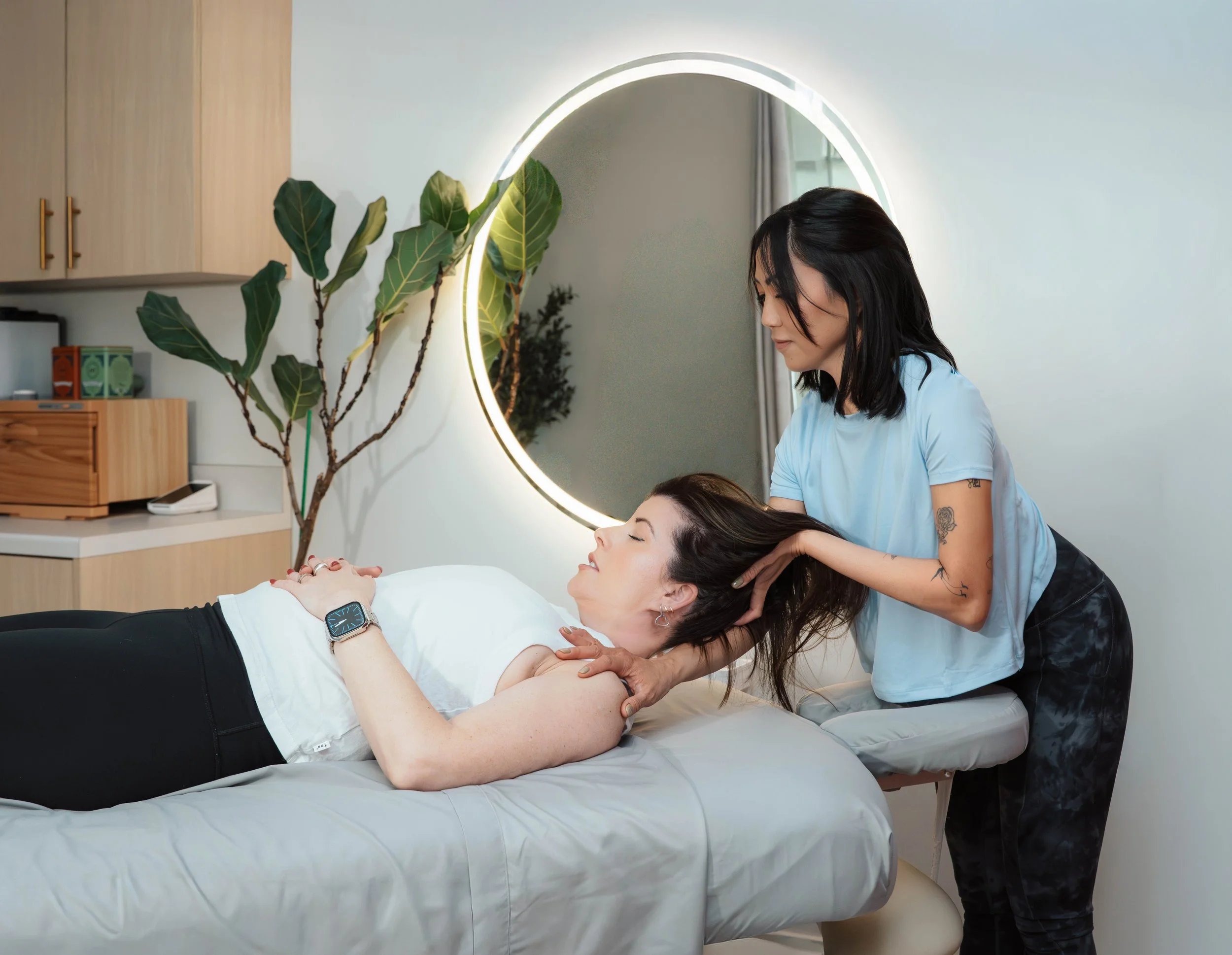 A woman receiving a massage from a masseuse in a modern, well-lit room with a large round mirror, a leafy plant, and wooden furniture.