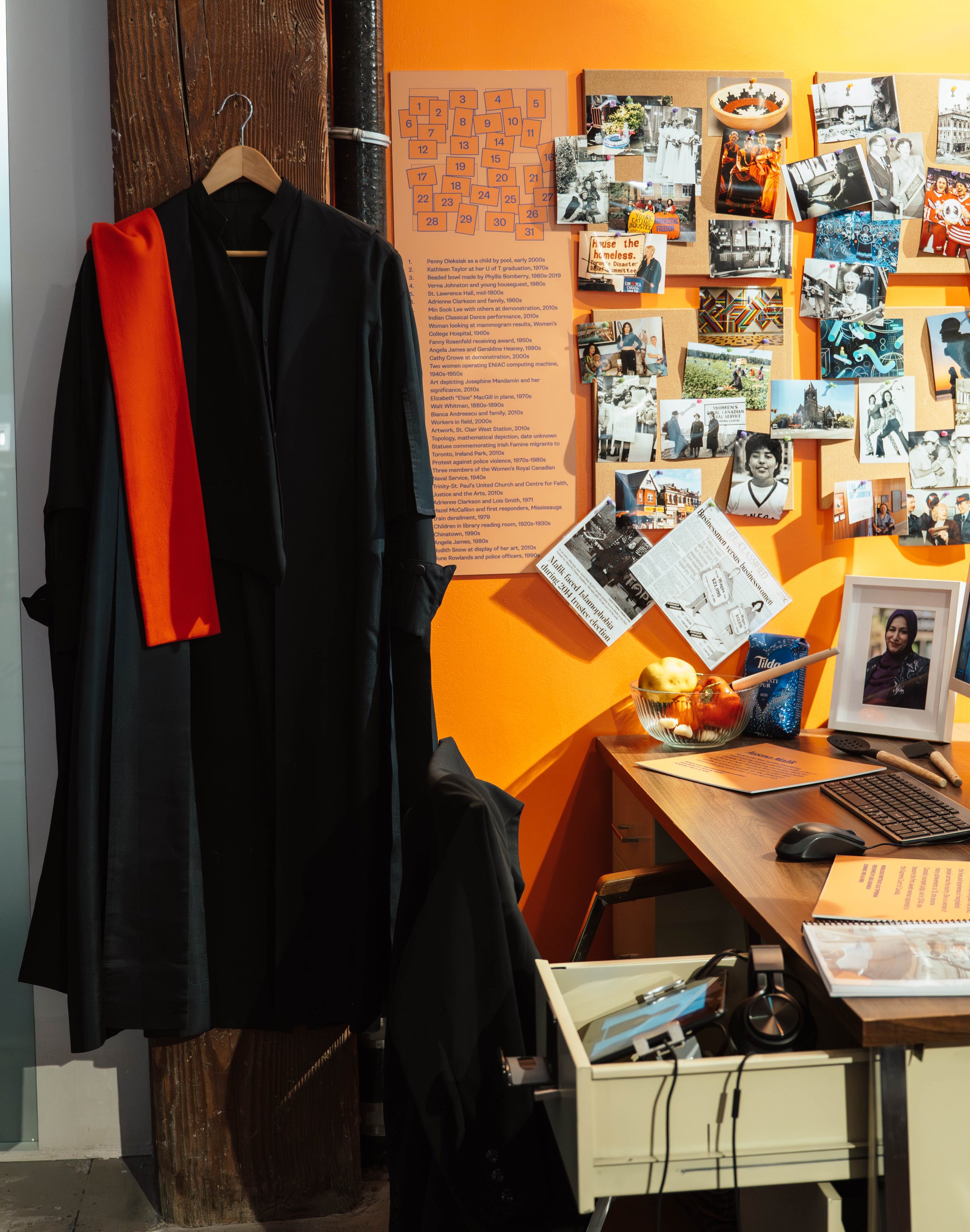An office or workspace with a mannequin dressed in a black gown with a red sash hanging on a hanger. The wall behind features photos, a crossword puzzle, and a newspaper collage on an orange background, as well as a desk with fruits, a framed photo o