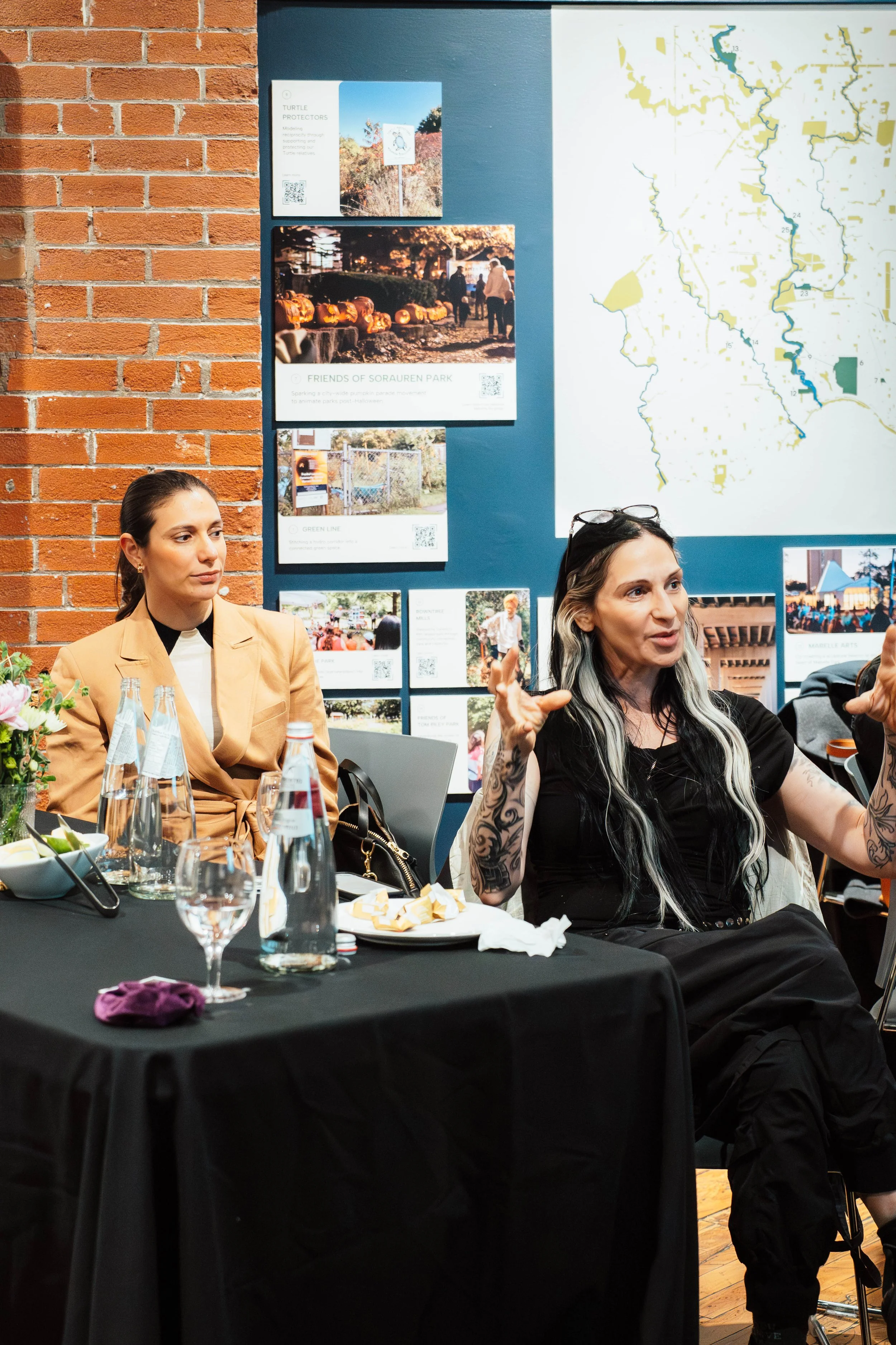 Two women sitting at a table during a discussion in a room with brick and blue walls, with posters and a map on the wall behind them.