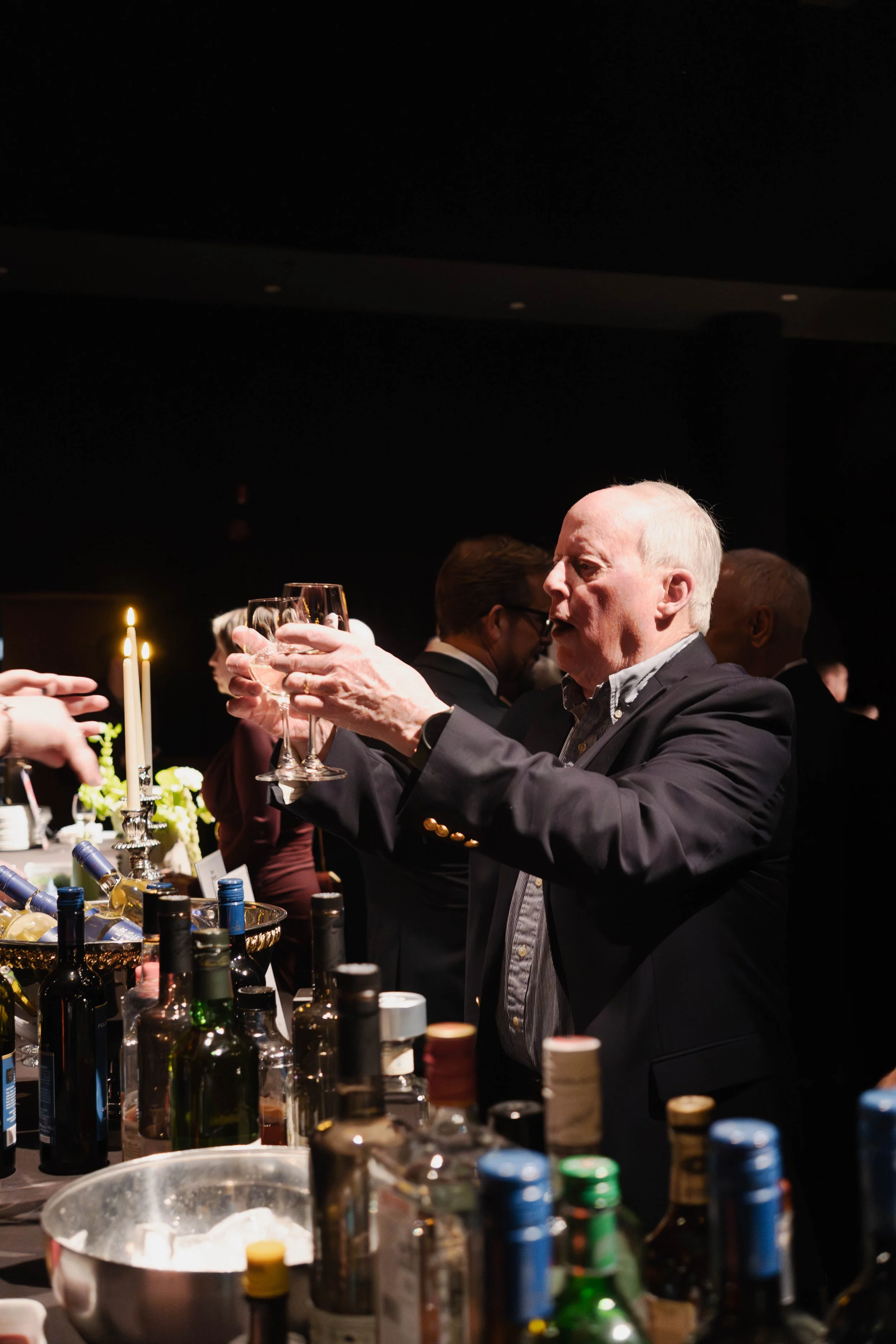 An elderly man in a dark suit raising a glass for a toast at a bar or party, with bottles and candles on the table.