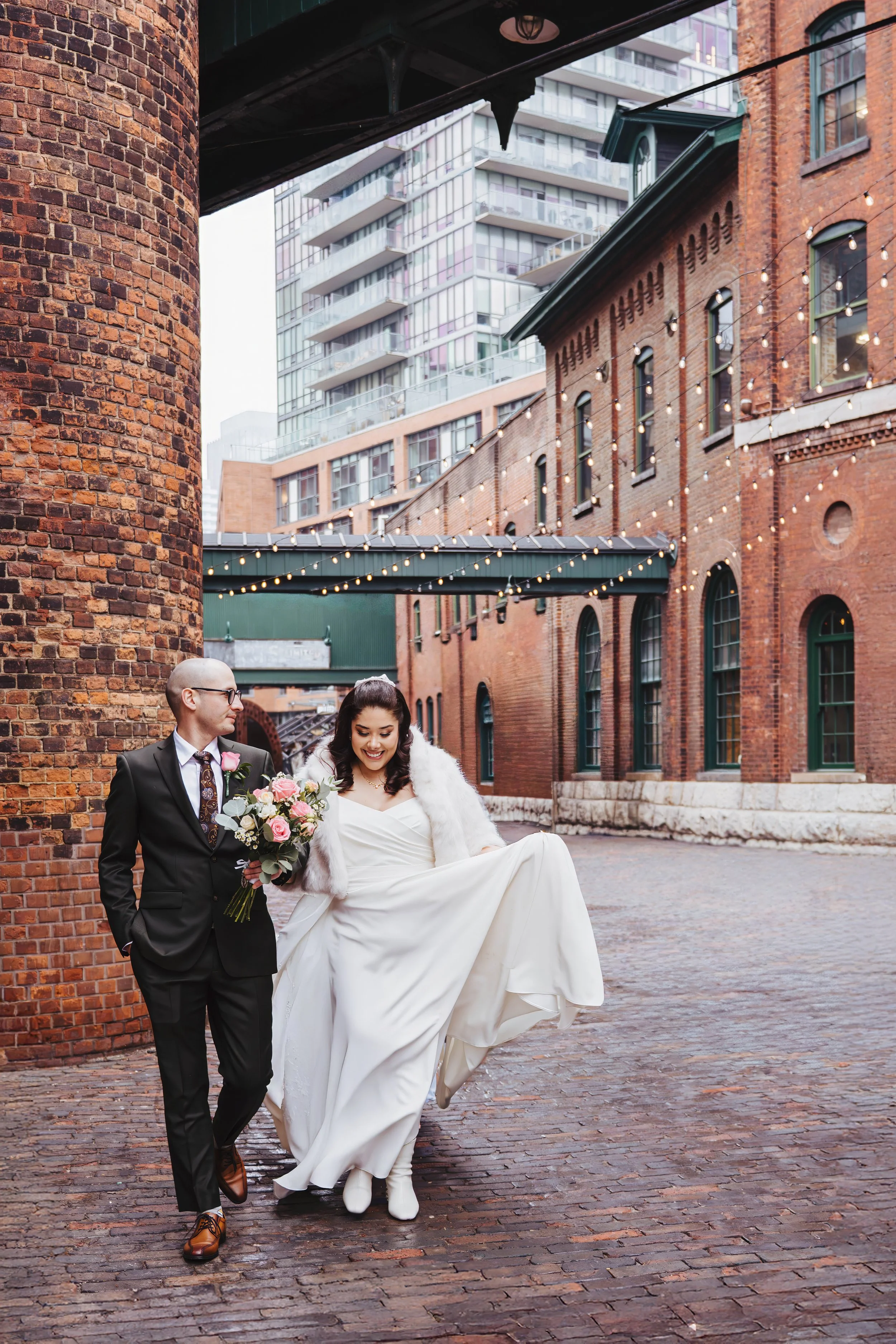 A bride and groom walking together outdoors in an urban area during daytime. The bride is wearing a white wedding dress and a white fur shawl, holding her dress up slightly, and smiling. The groom is dressed in a black suit with a white shirt, patter