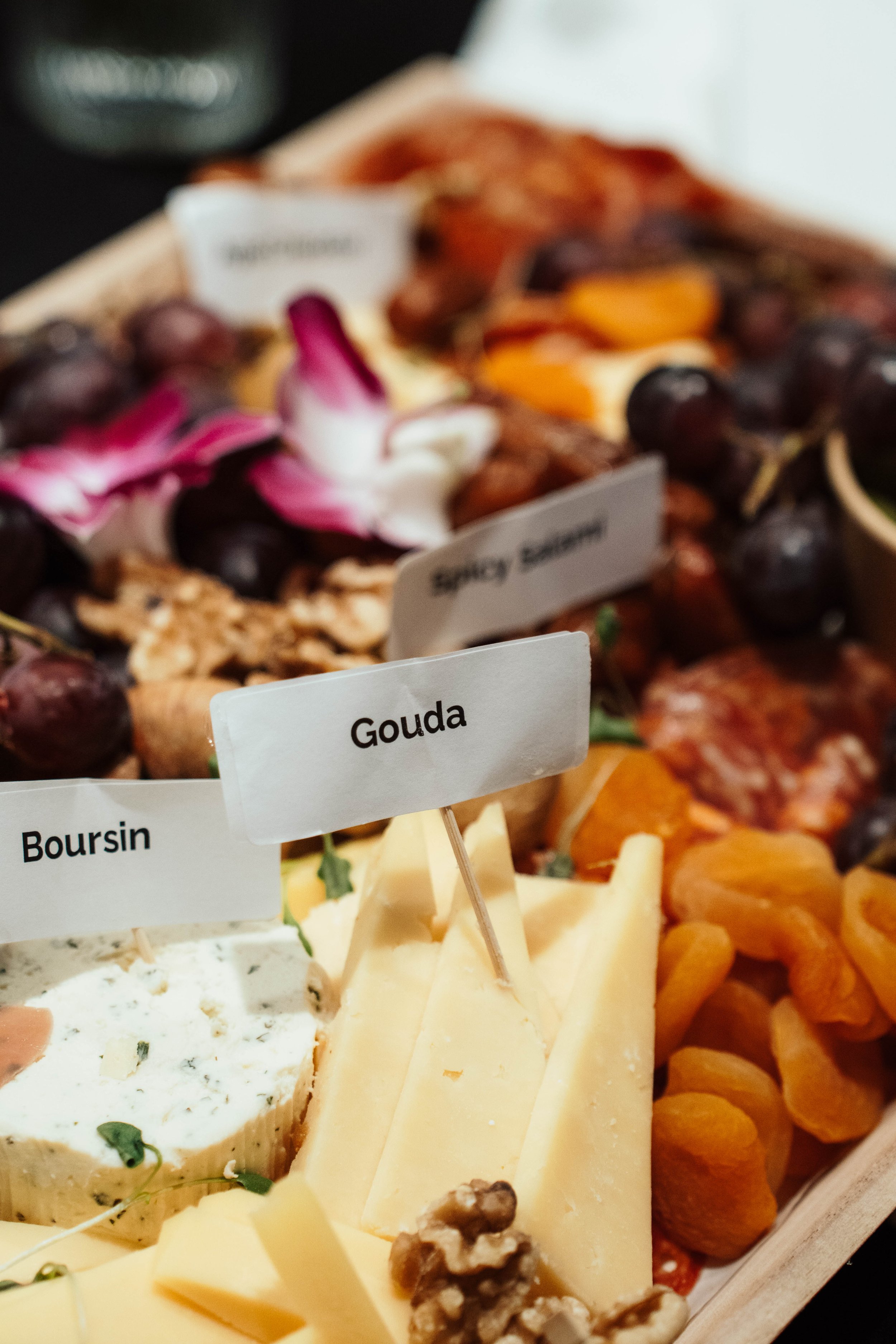 Assorted cheeses including Gouda, Boursin, and Blueberry Bourbon, garnished with dried apricots, grapes, nuts, and edible flowers on a cheese platter.