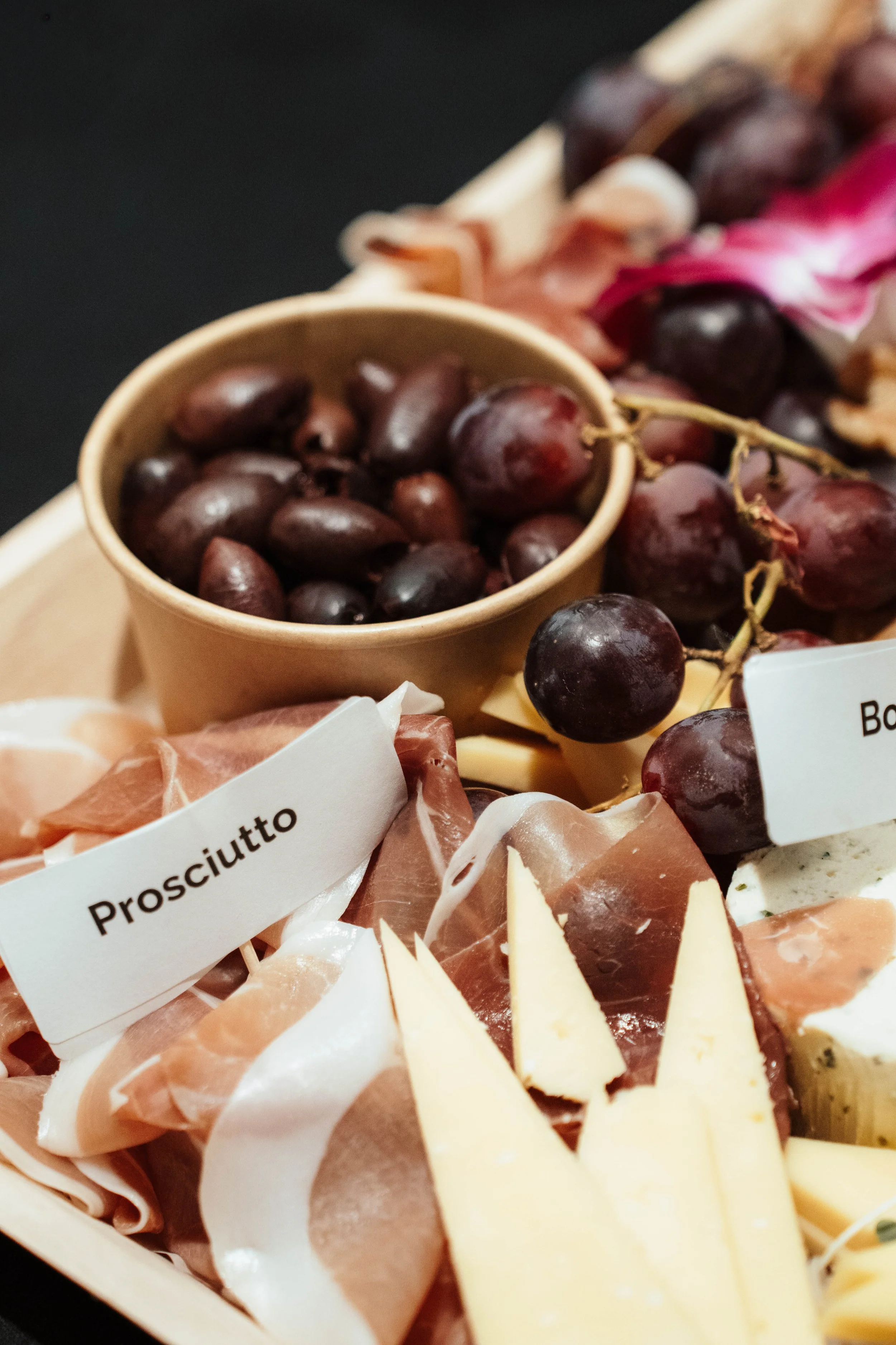 A cheese and charcuterie board with grapes, prosciutto, and other cheeses, labeled with tags for prosciutto and bordeaux grapes.