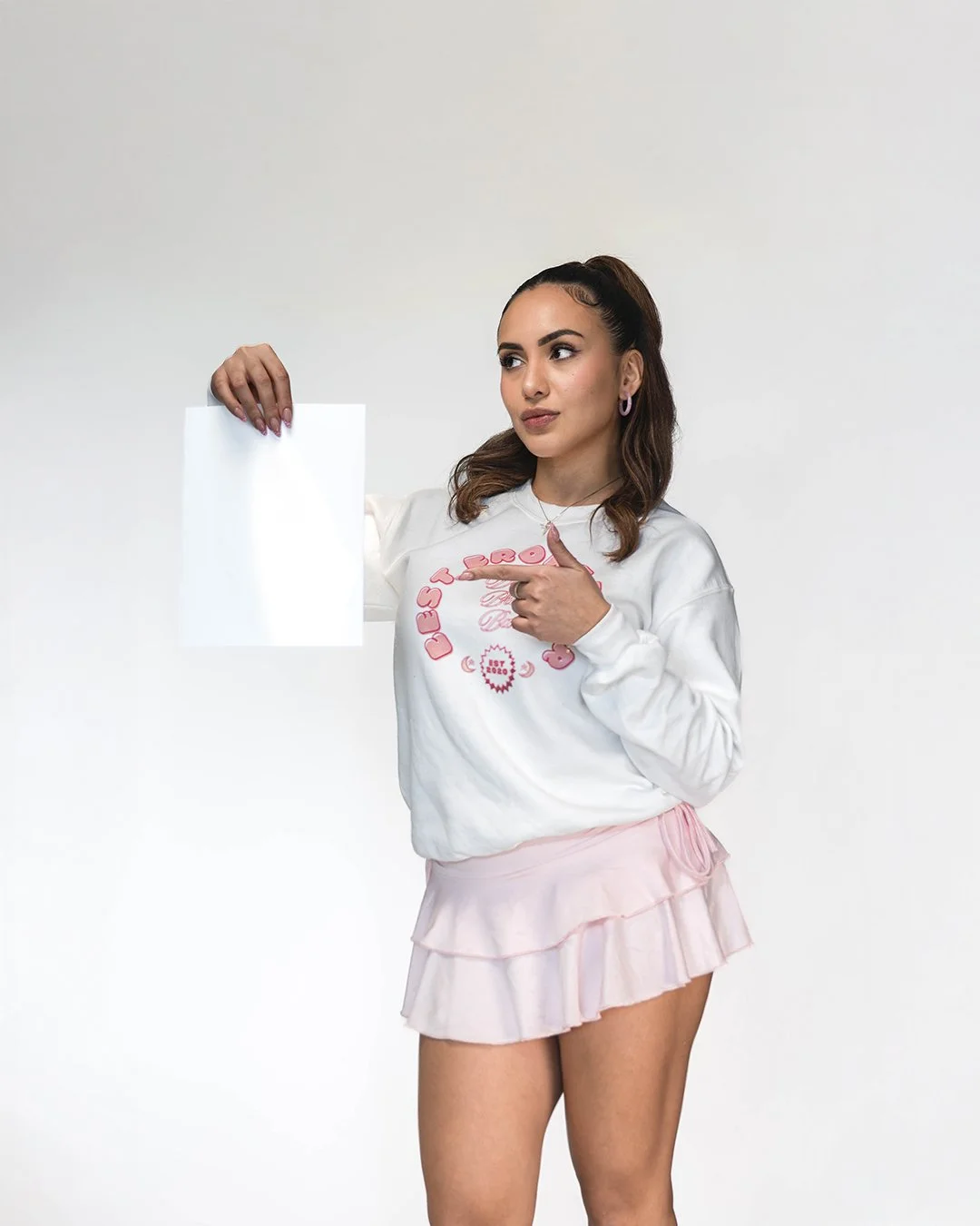 A young woman with brown hair in a high ponytail, wearing a white sweatshirt with pink graphics and lettering, and a light pink ruffled skirt, points at an empty white sheet of paper she holds up against a plain white background.