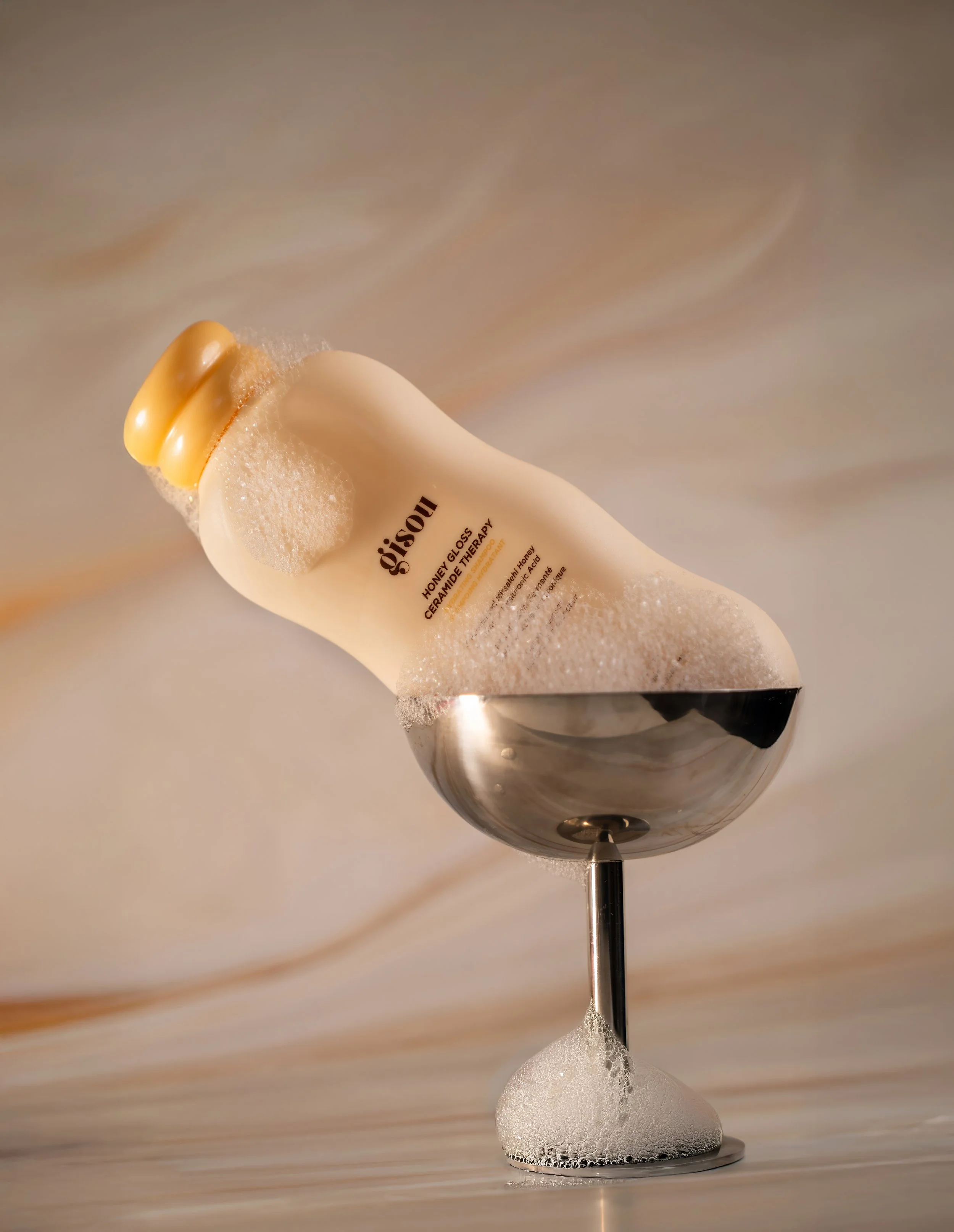 A bottle of honey gloss balm leaning against a metal scoop filled with soapy foam, with frothy bubbles on the bottle and at the base of the scoop.