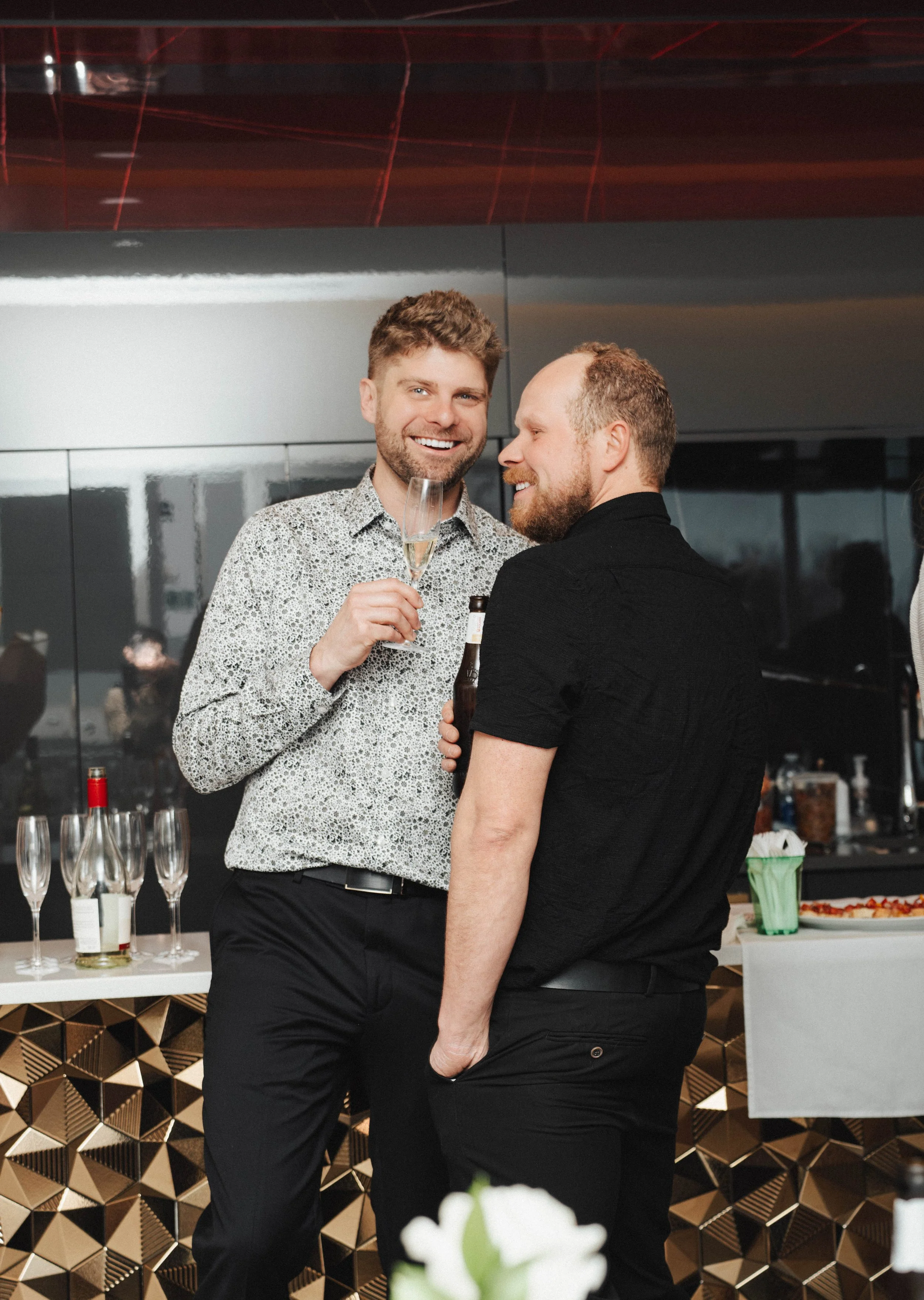 Two men are at a social gathering, one is smiling and holding a champagne flute, and the other is leaning in close, holding a beer bottle. They are standing near a bar with champagne glasses and bottles, and colorful food is visible in the background