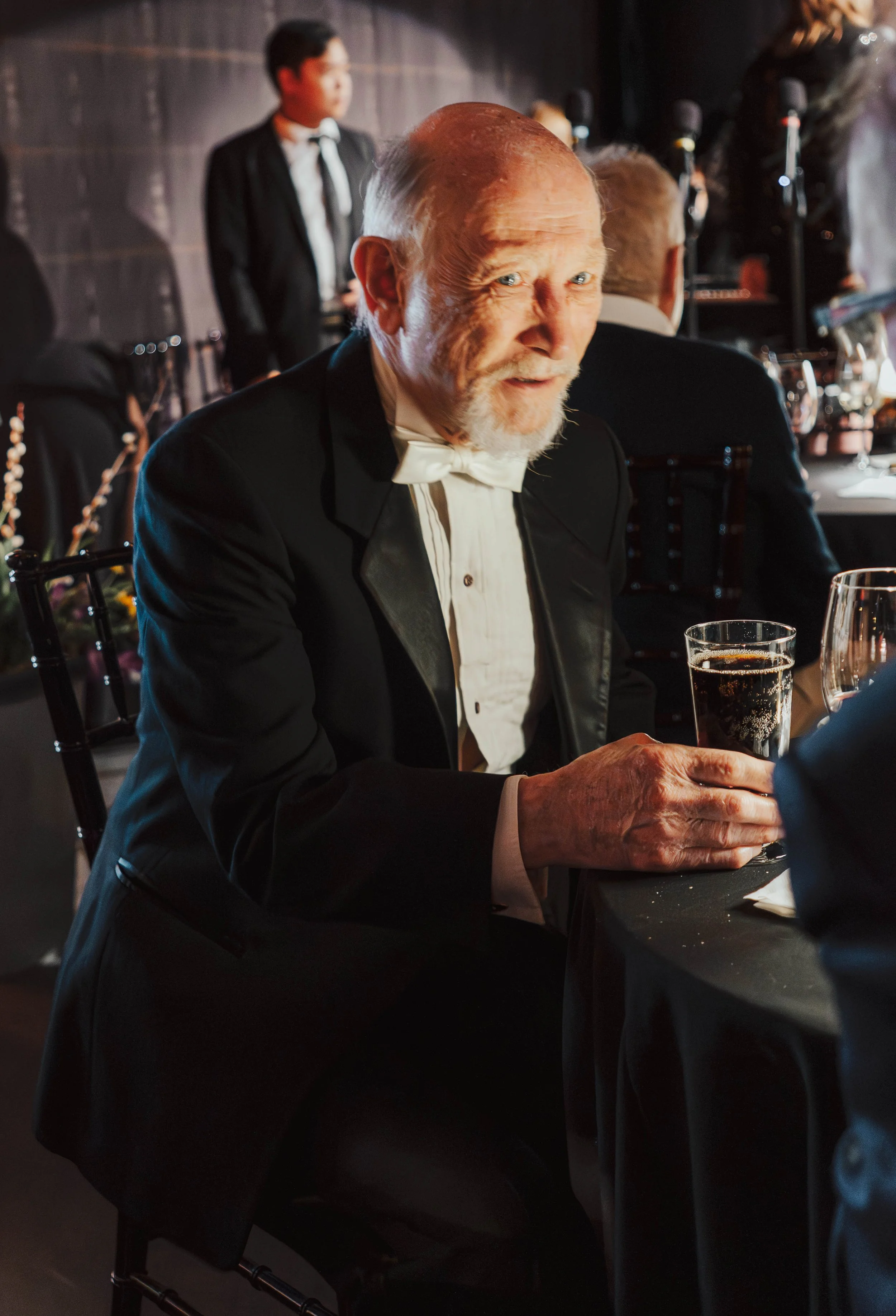 An elderly man with a white beard and mustache, dressed in a tuxedo with a bowtie, sitting at a formal event table, holding a glass of dark soda, and smiling at the camera.