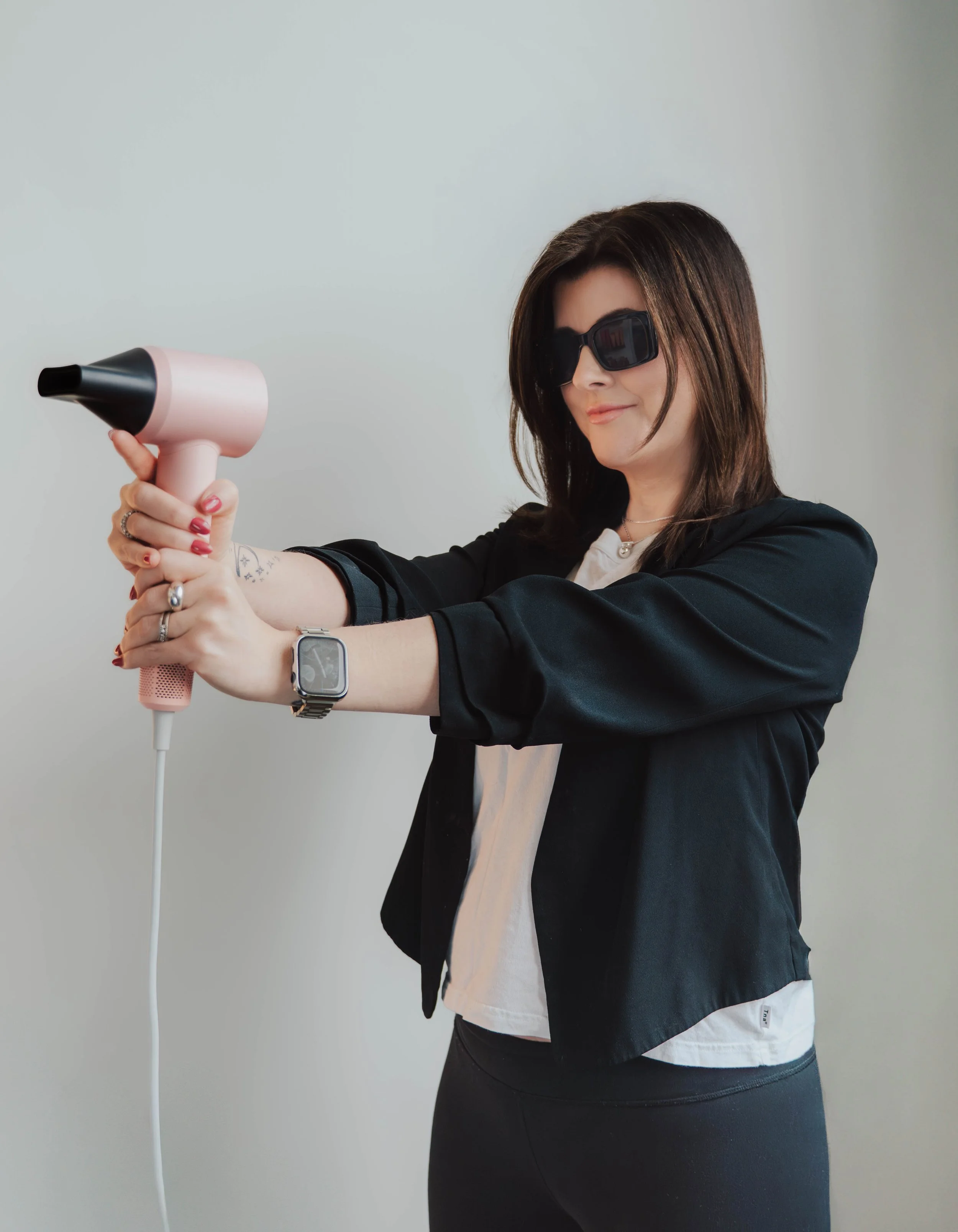 Woman wearing sunglasses using a pink hairdryer with a black nozzle.