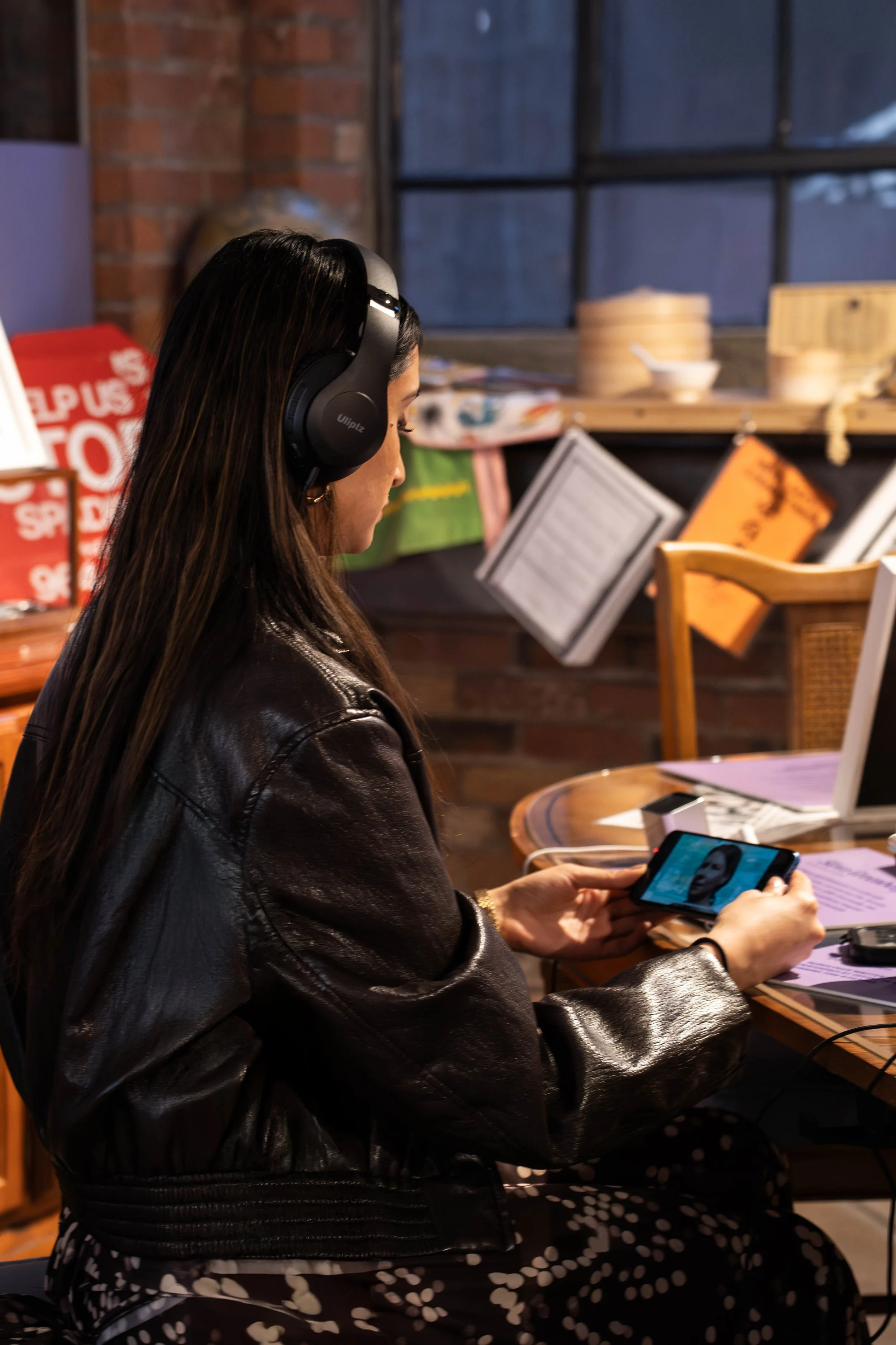 A woman with long dark hair wearing black headphones and a leather jacket, sitting at a table, looking at her smartphone in a cafe with a brick wall and shelves with bowls and paper menus in the background.