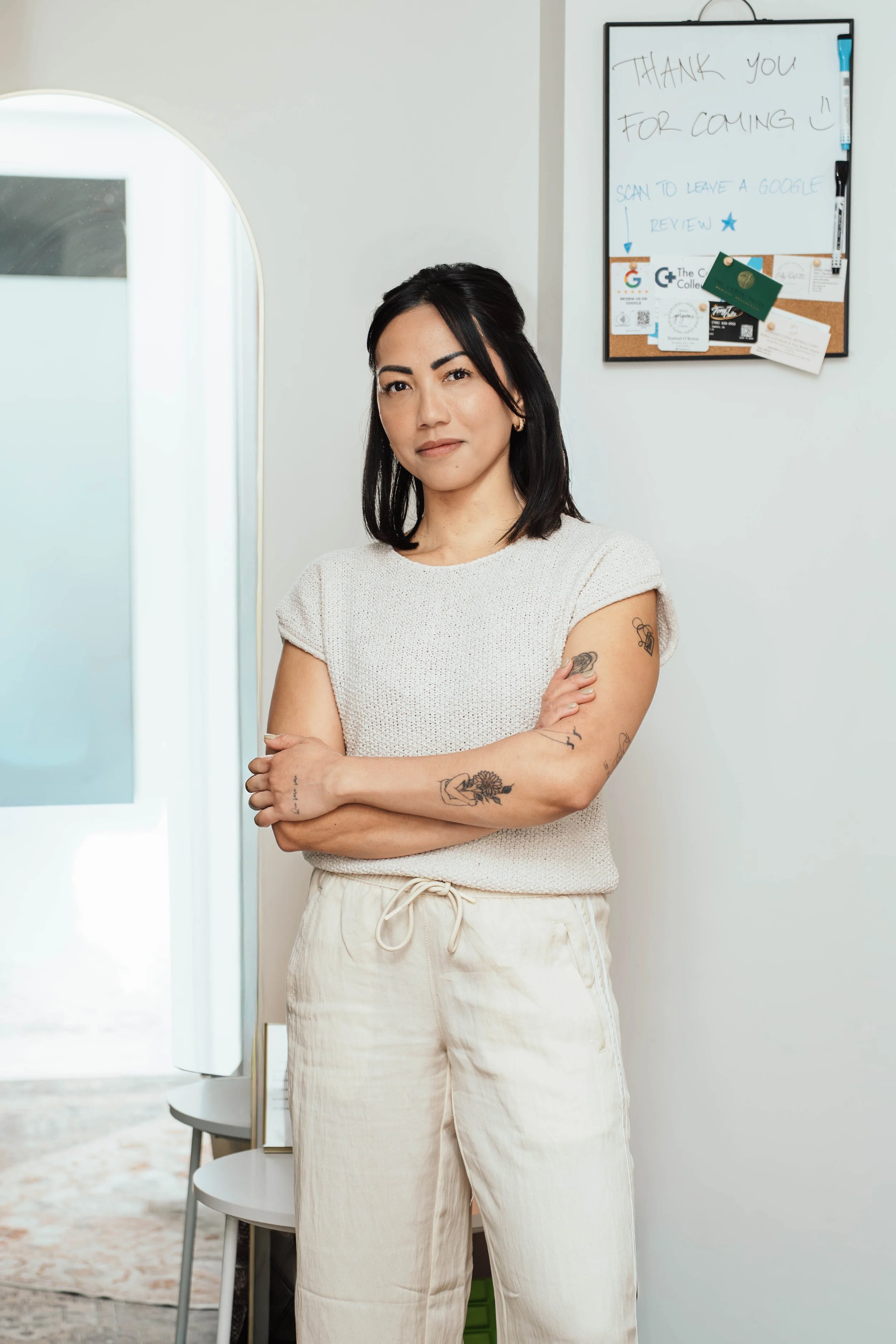 A woman with shoulder-length black hair, tattoos on her left arm, wearing a white sleeveless top and cream-colored pants, standing with arms crossed in front of a white wall with a whiteboard that has a handwritten message.