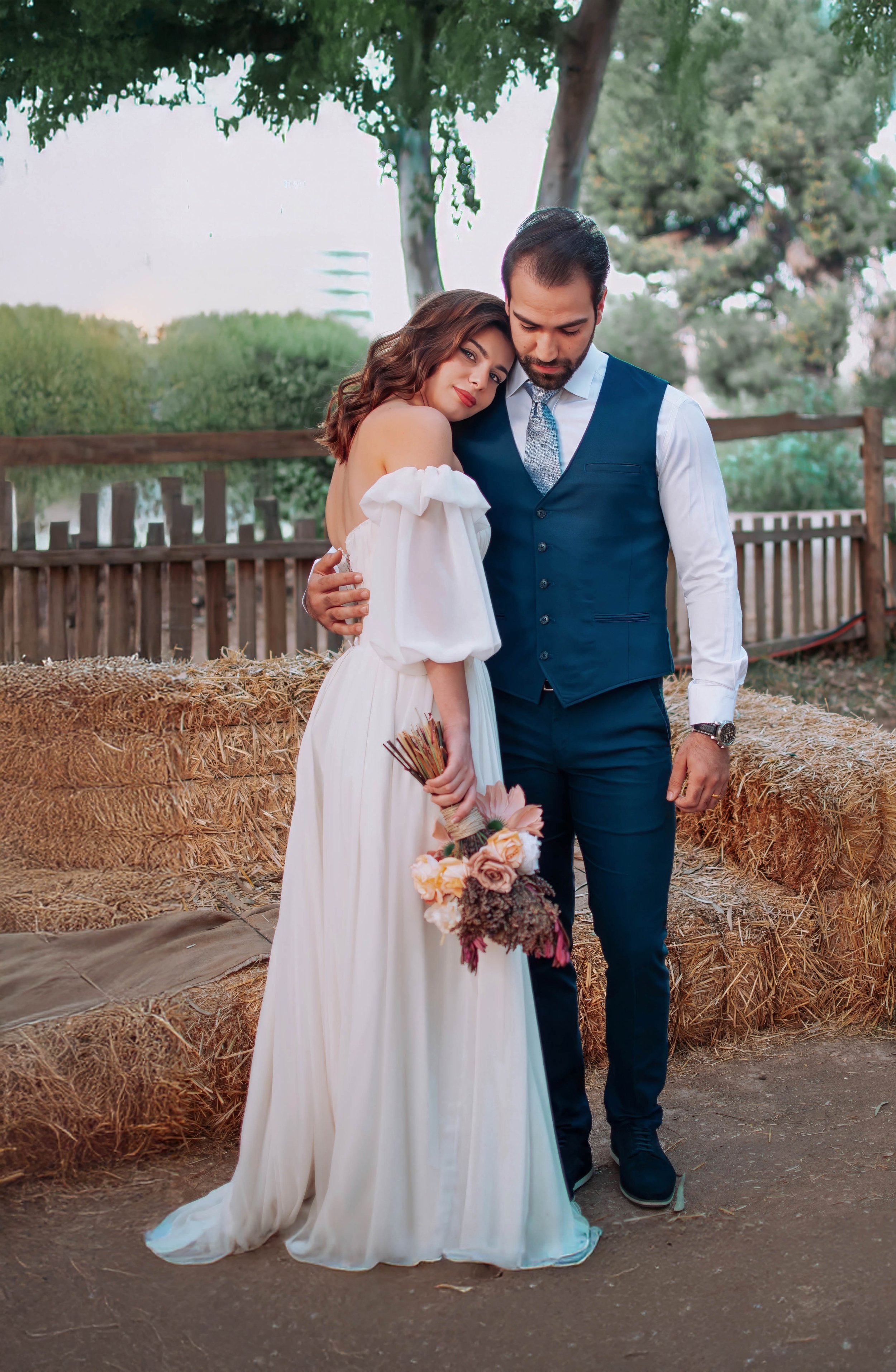 A couple dressed in wedding attire, the woman holding a bouquet, embracing outdoors near hay bales and a wooden fence, with trees in the background.