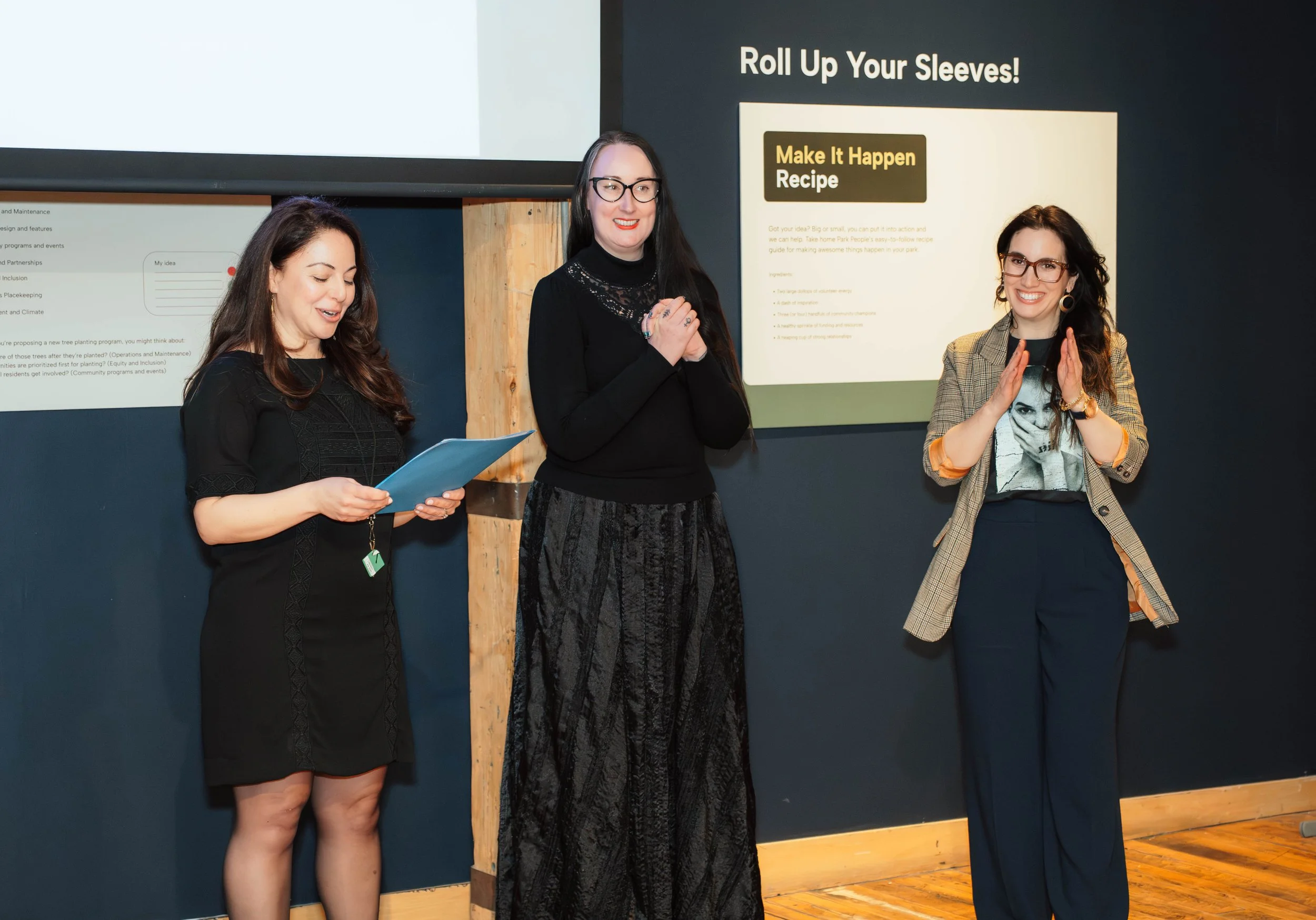 Three women standing in front of a presentation screen in a room with wooden floor. One woman on the left is reading from a folder, the woman in the middle is smiling with her hands clasped, and the woman on the right is clapping. The screen displays