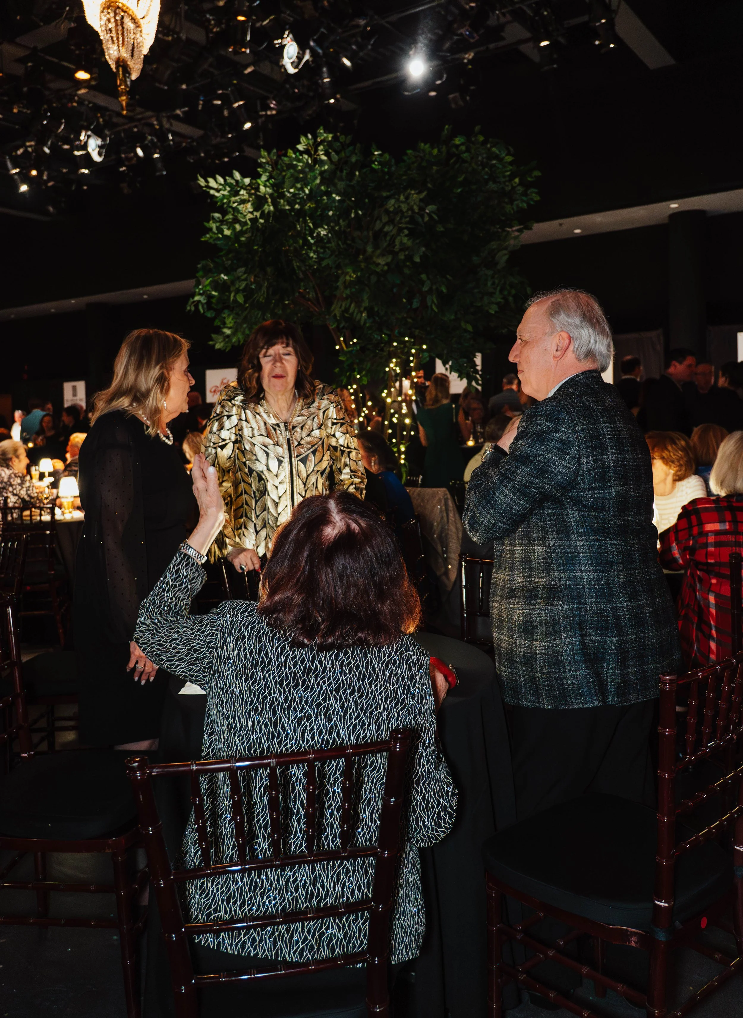 People having a lively conversation at an elegant indoor event, with a large green tree decorated with lights in the background.