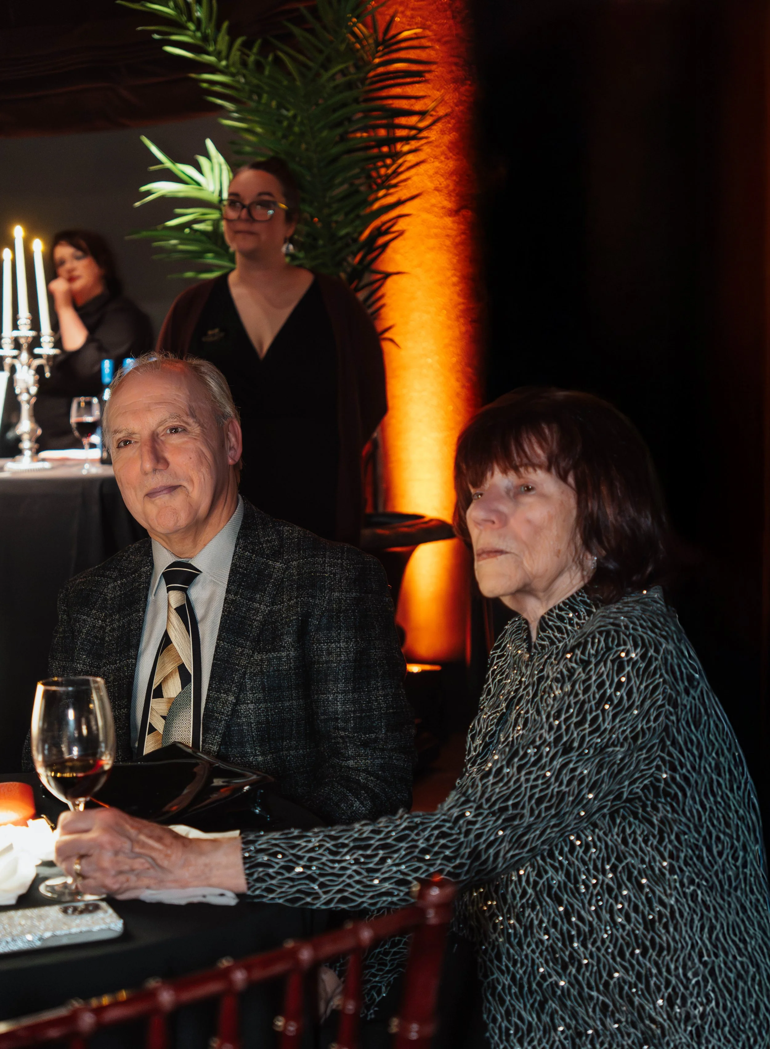 An elderly man and woman seated at a dinner event, with a candle-lit table in front of them and a large green plant and warm orange lighting in the background. A woman with glasses stands in the background.