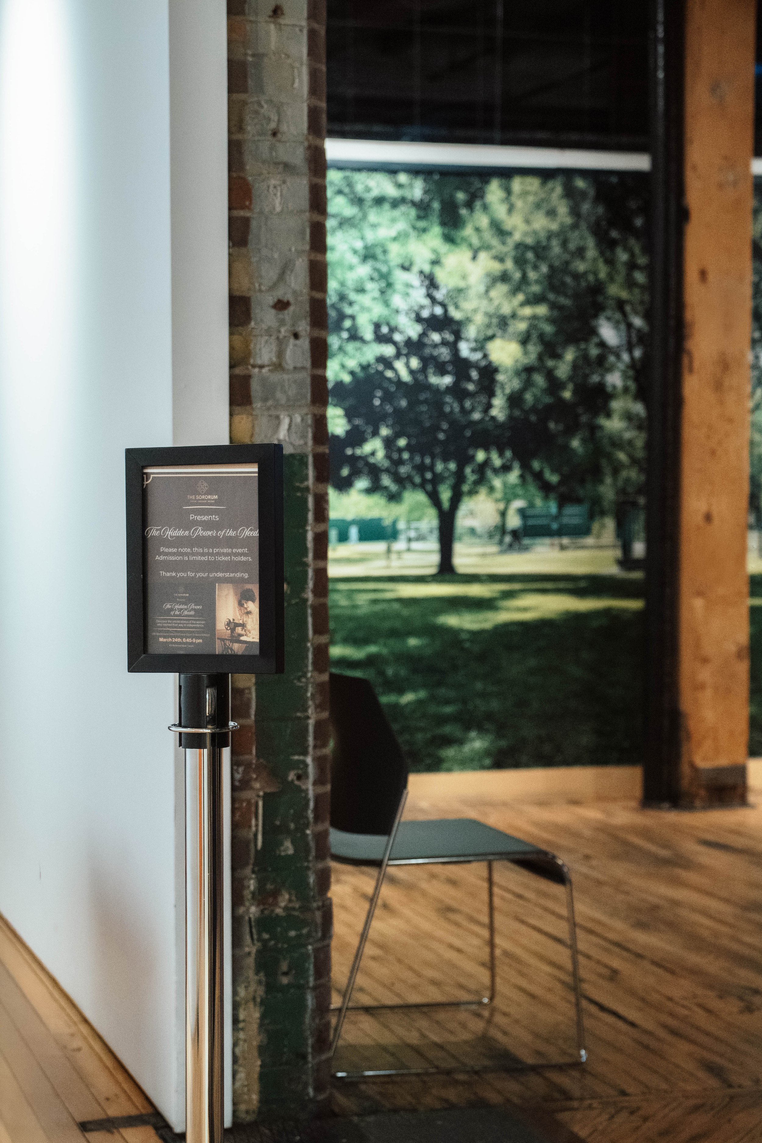 Interior view of a building with a sign on a stand and a black chair. The background shows a large window with a view of a park with trees and benches.