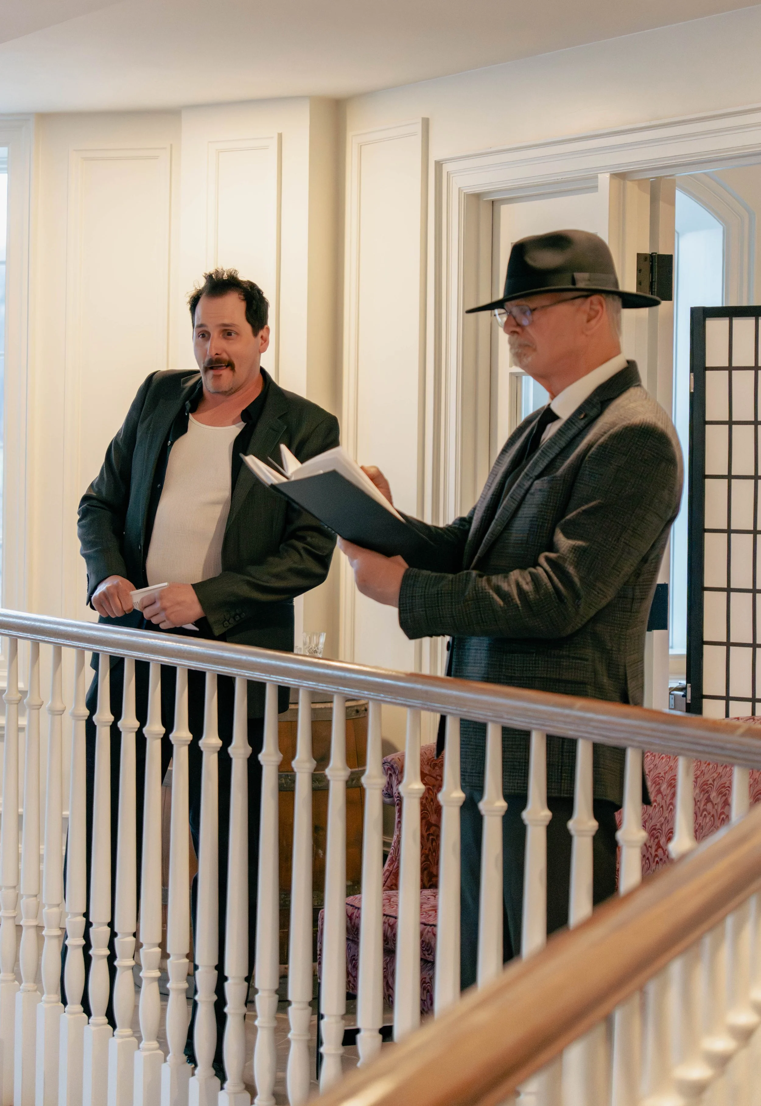 Two men standing on a staircase railing inside a house. One man is holding a book, and the other is reading from a notebook. The man reading is wearing a gray checked blazer, black hat, glasses, and a white shirt. The other man is wearing a black bla
