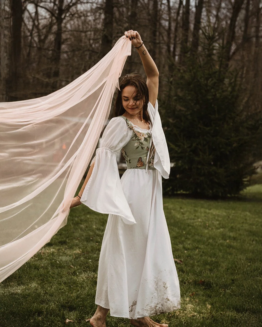 A woman standing outdoors on grass, holding a sheer fabric runway above her head. She is wearing a white skirt, a floral top, and a white jacket, with a tattoo on her arm and barefoot. Looks relaxed with a forest background.