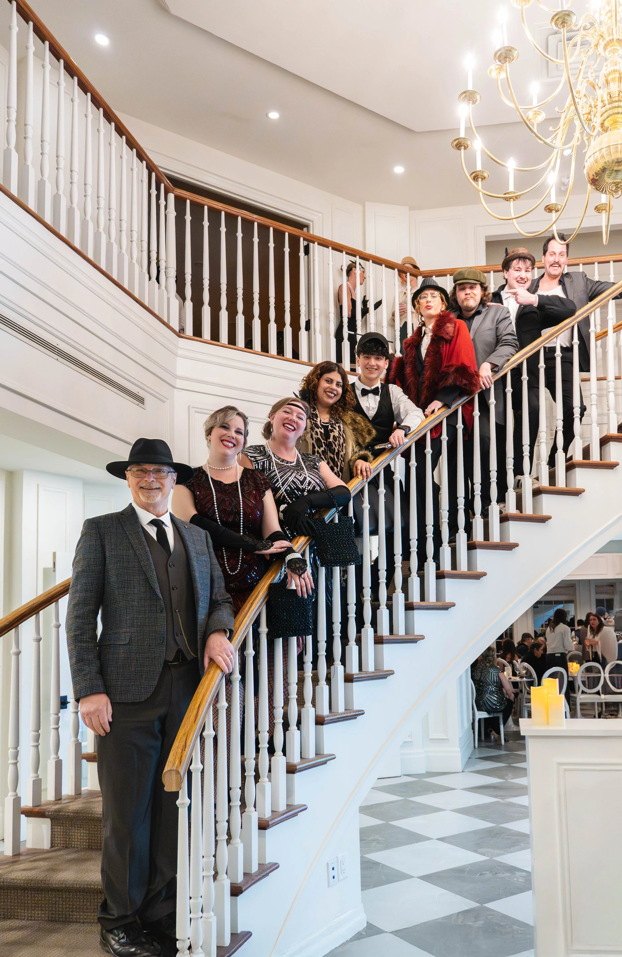 Group of people dressed in vintage and formal attire standing on a curved staircase inside a well-lit elegant venue with guests dining below.