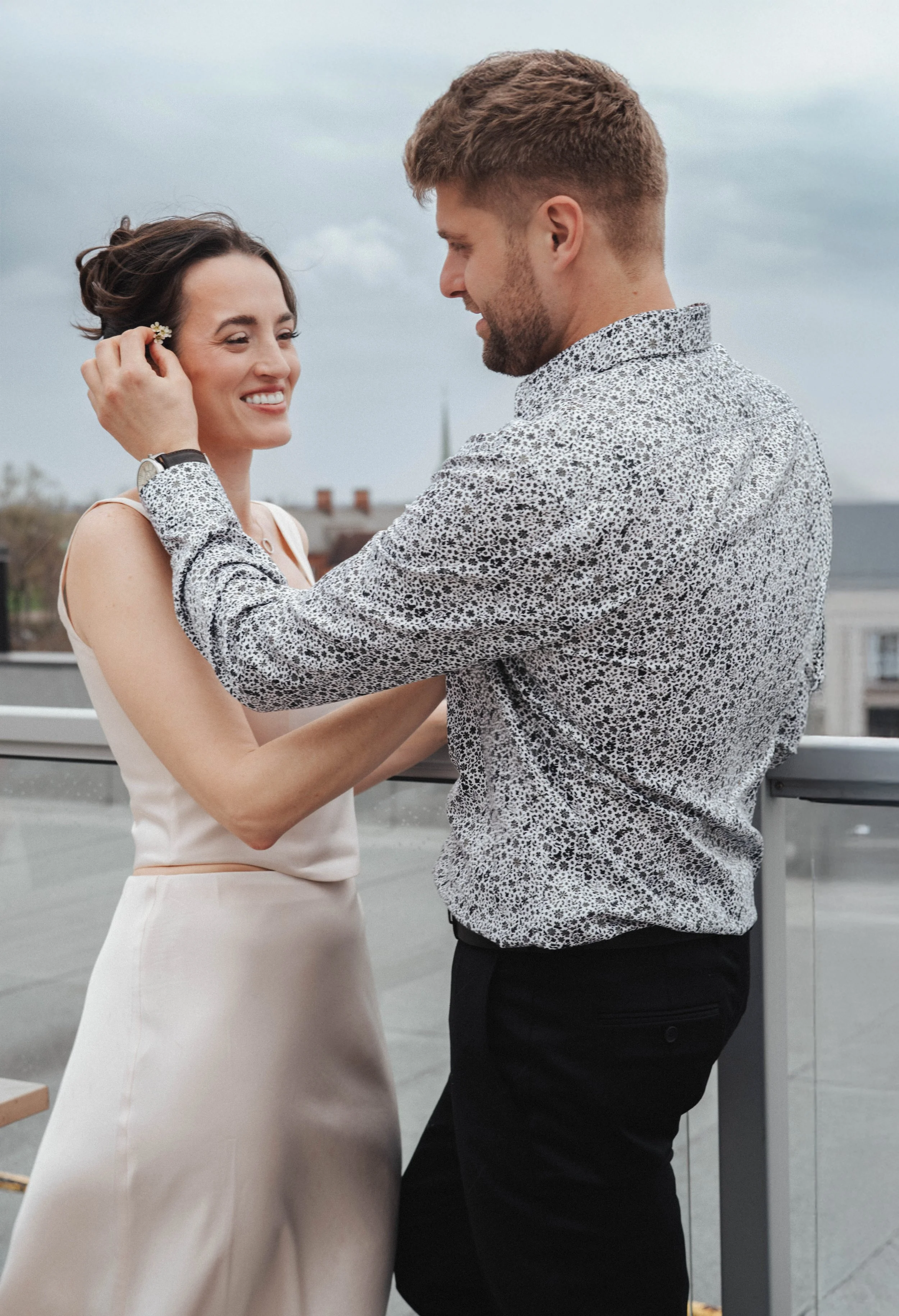 A man and a woman are holding hands in an outdoor setting, looking at each other, with overcast skies in the background.