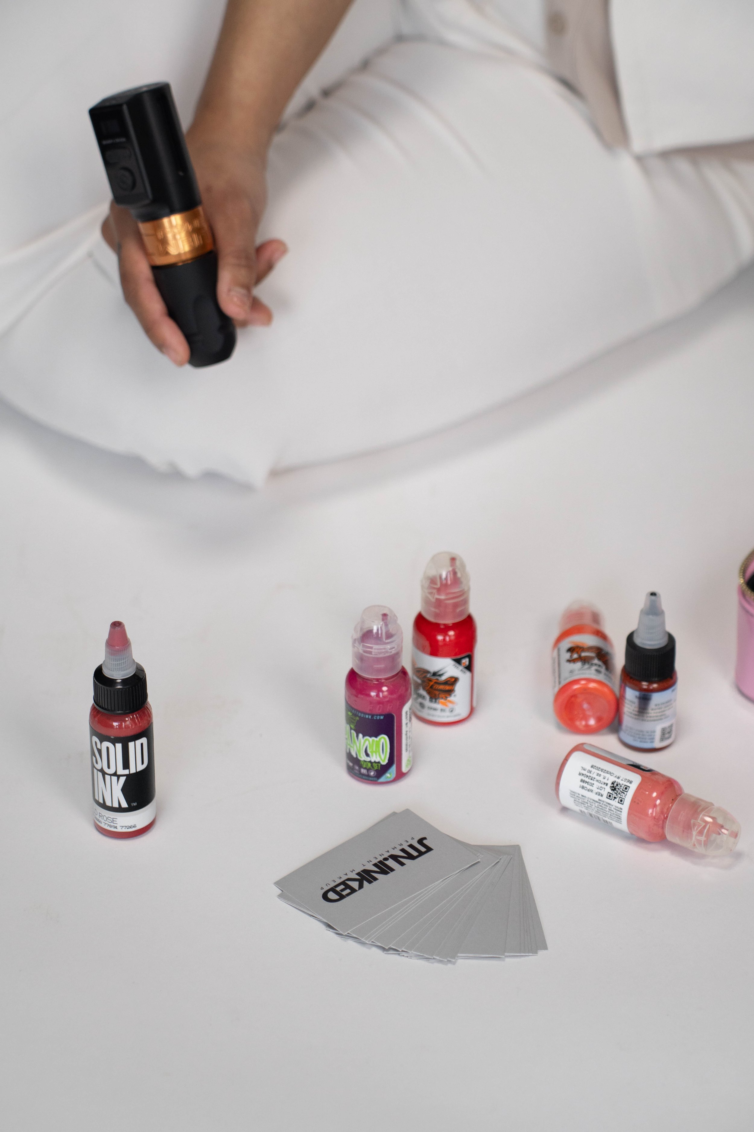 Person holding a tattoo gun near tattoo ink bottles and a deck of tattoo-related playing cards on a white table.