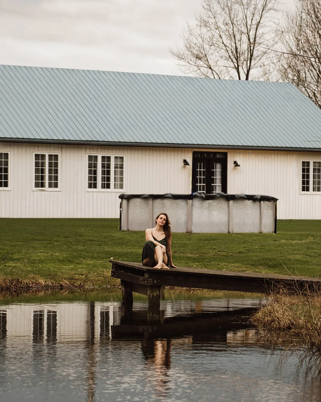 A woman sitting on a small wooden dock by a pond with a large white house and a metal hot tub in the background.