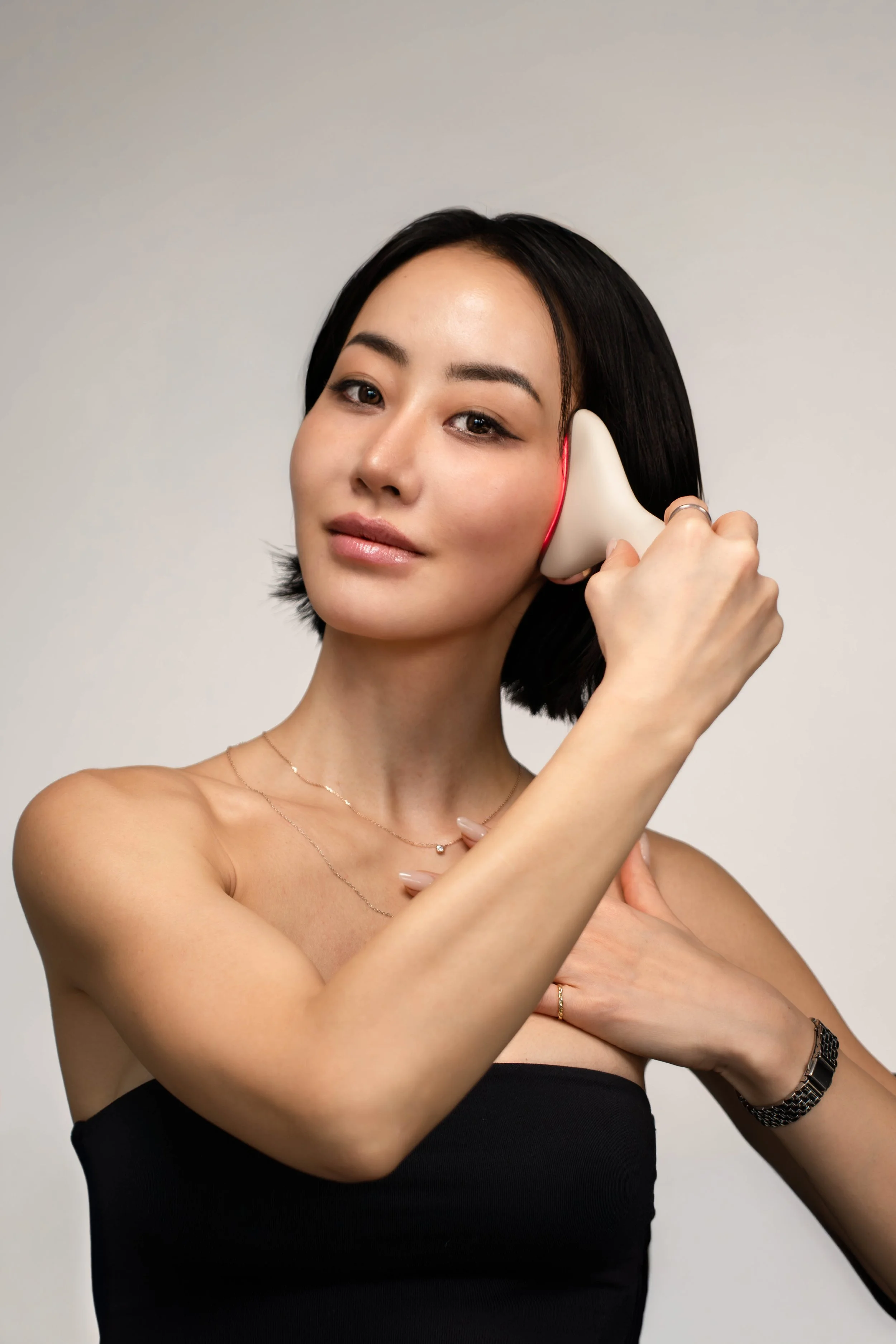 A woman holding a facial massager to her face, looking at the camera, wearing a black strapless top and jewelry against a plain background.