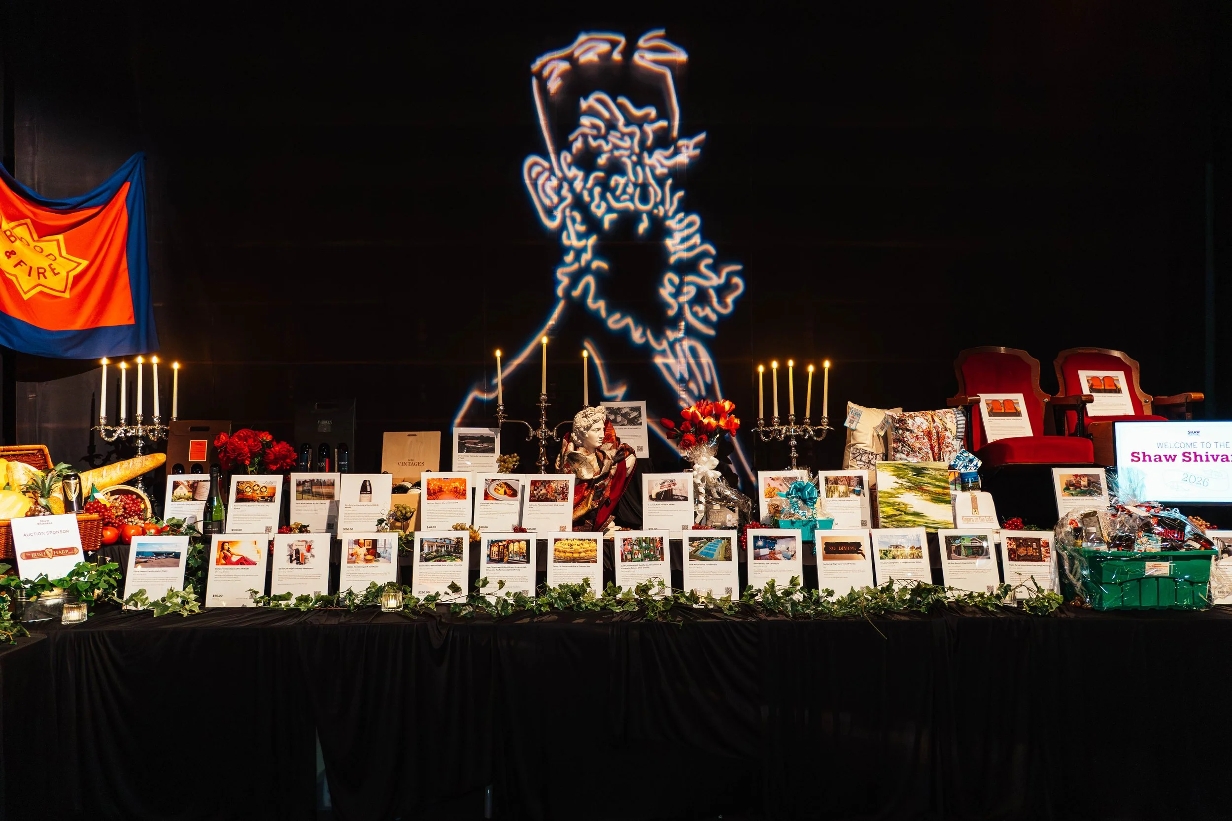 A display table at an auction event with various items, photographs, flower arrangements, and candelabras. A neon outline of Abraham Lincoln's face is on the black wall behind the table. On the left, a flag with a fire badge is hanging. On the right,