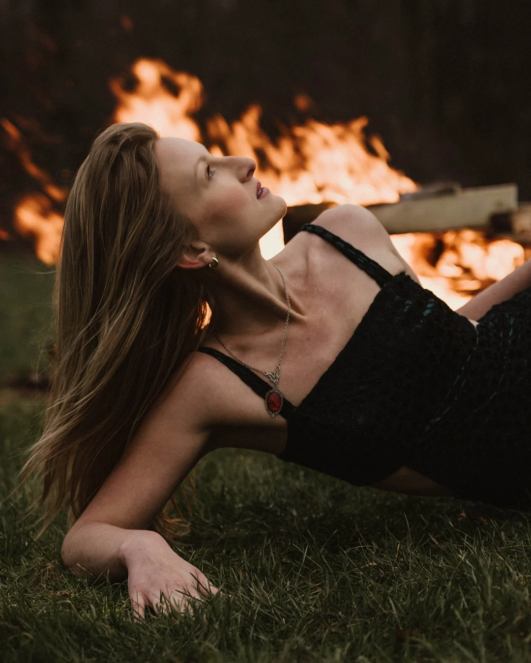 A woman with long blond hair lying on the grass with a fire burning in the background.