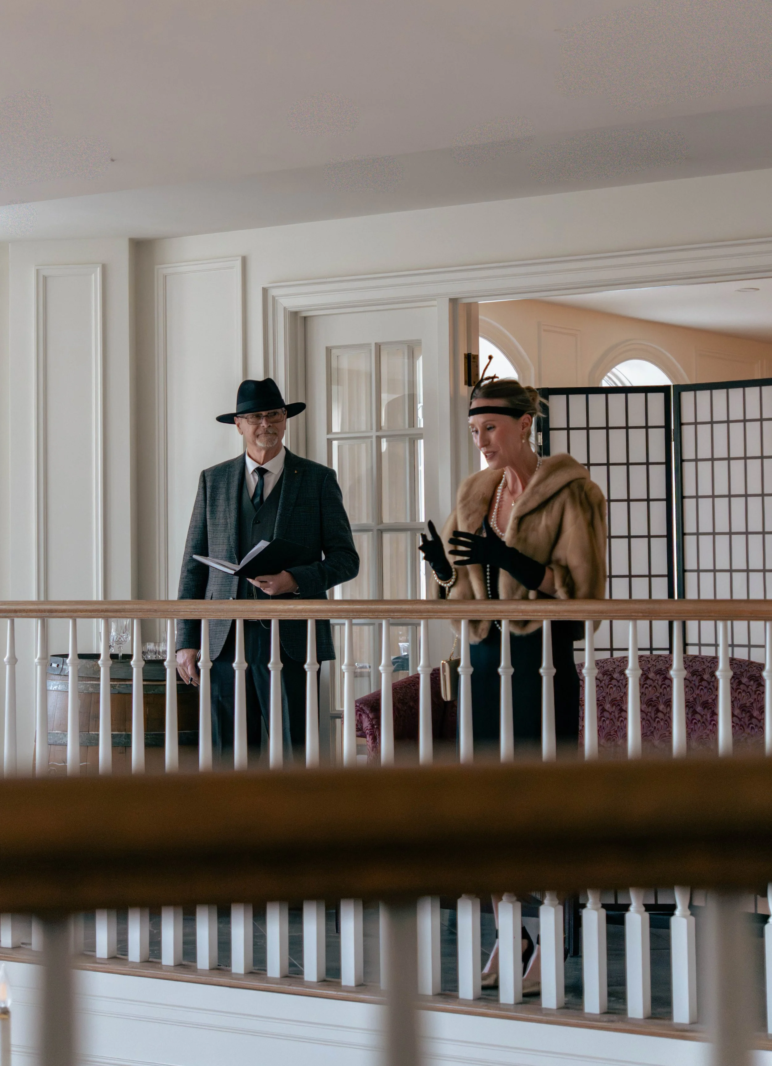 A man dressed in a vintage-style suit with a fedora hat and glasses holds a book, standing next to a woman wearing a fur stole, pearl jewelry, black gloves, and a vintage headband, engaging in conversation in a brightly lit indoor setting with white 