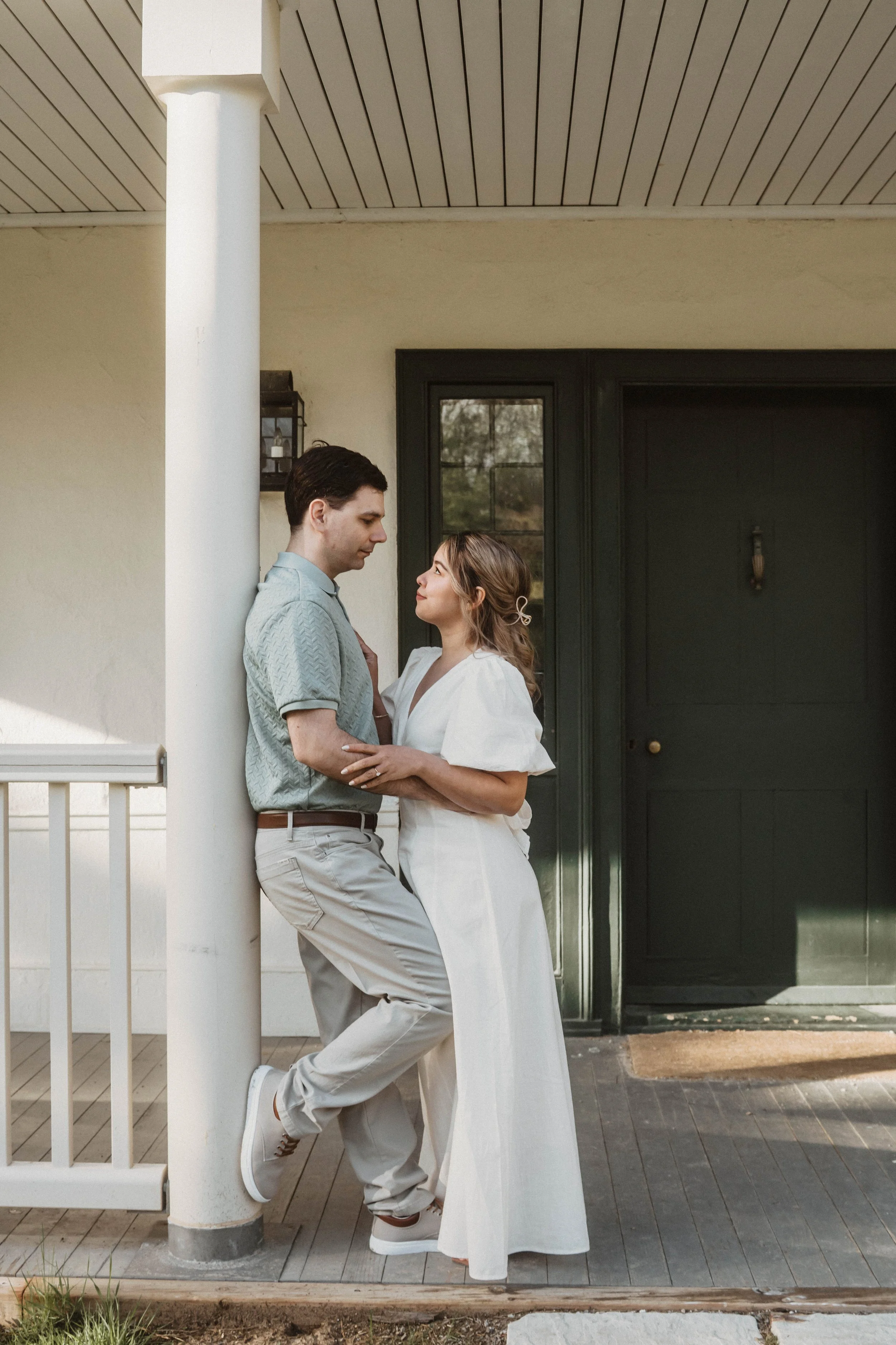 A man and woman standing close to each other on a porch in front of a dark green door, with the man leaning against a white pillar and looking at the woman, and the woman gazing back at him.