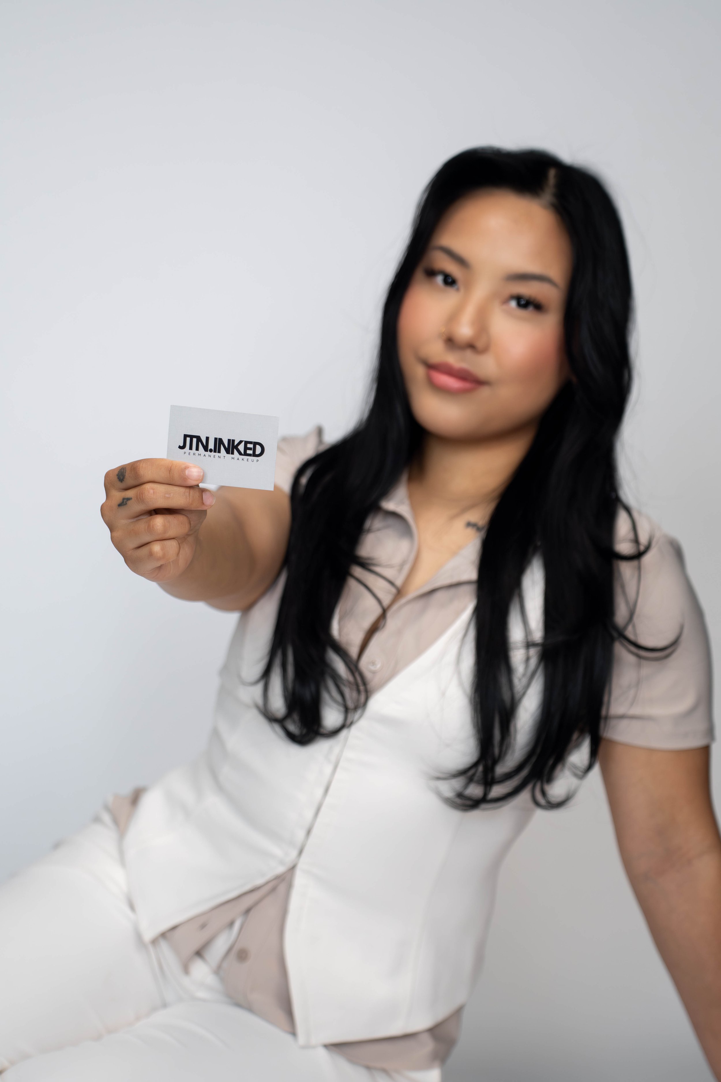A woman with long black hair holding a business card that says 'JIN.NKED PERMANENT MAKEUP' in front of a plain white background.