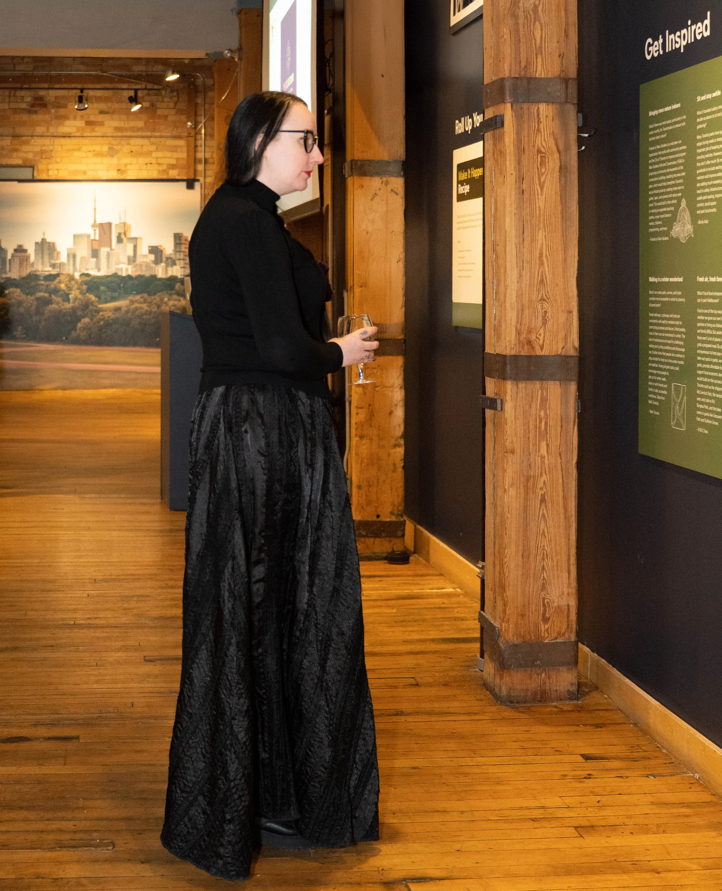 A woman with black hair, glasses, and dressed in black top and long black patterned skirt, stands in an art gallery or exhibit space, holding a glass of wine and looking at informational posters on the wall.