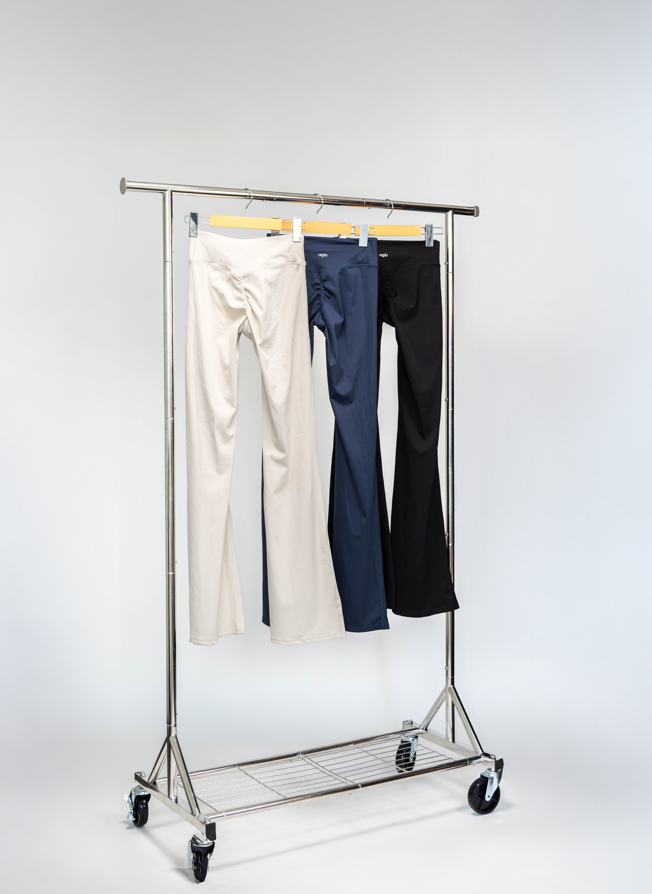Three pairs of pants hanging on a metal clothing rack with wheels, against a plain white background. The pants are in cream, navy blue, and black colors.