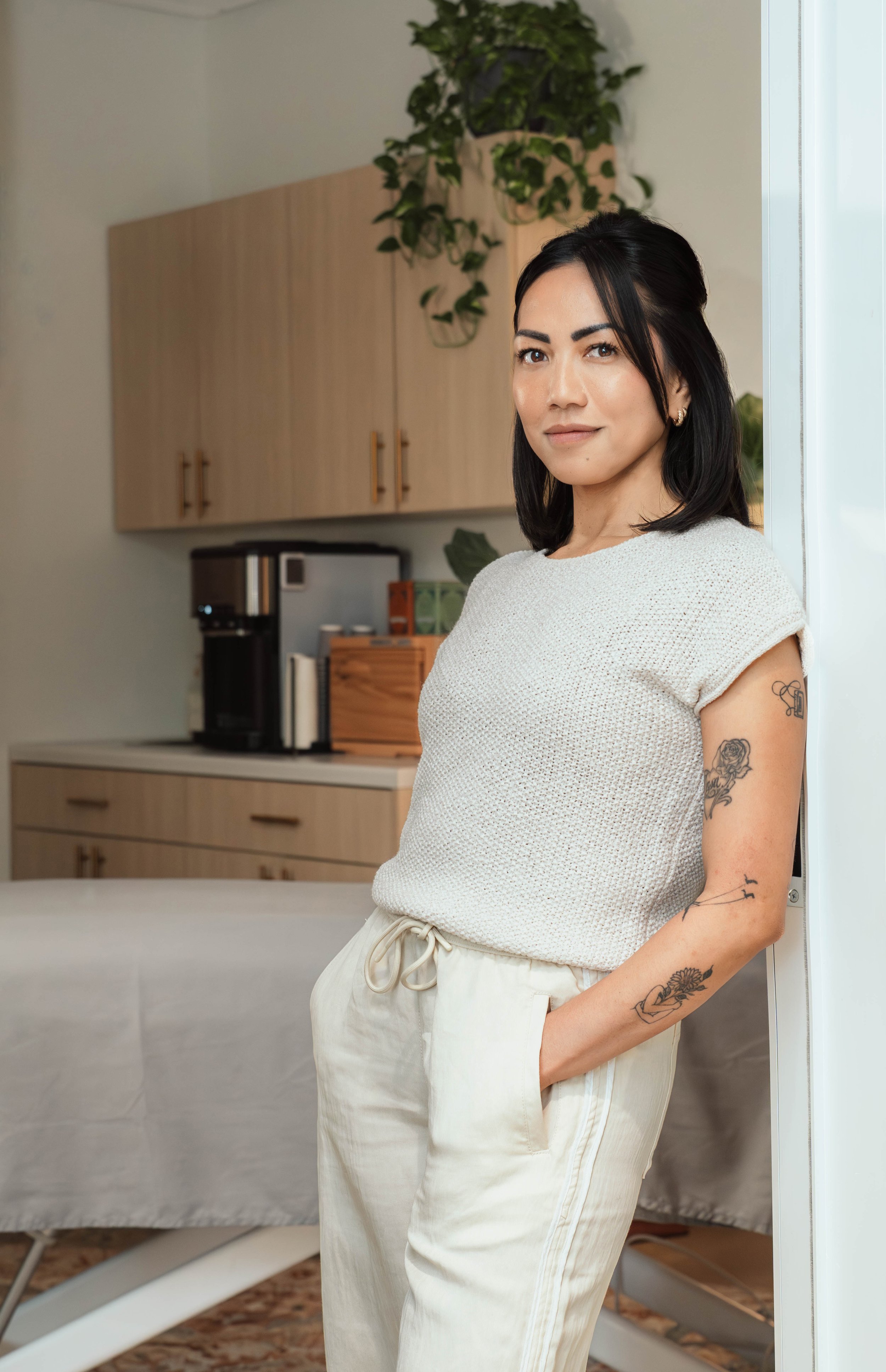 A woman standing indoors near a wall, wearing a white knit top and light-colored pants, with black hair and tattoos on her arm.