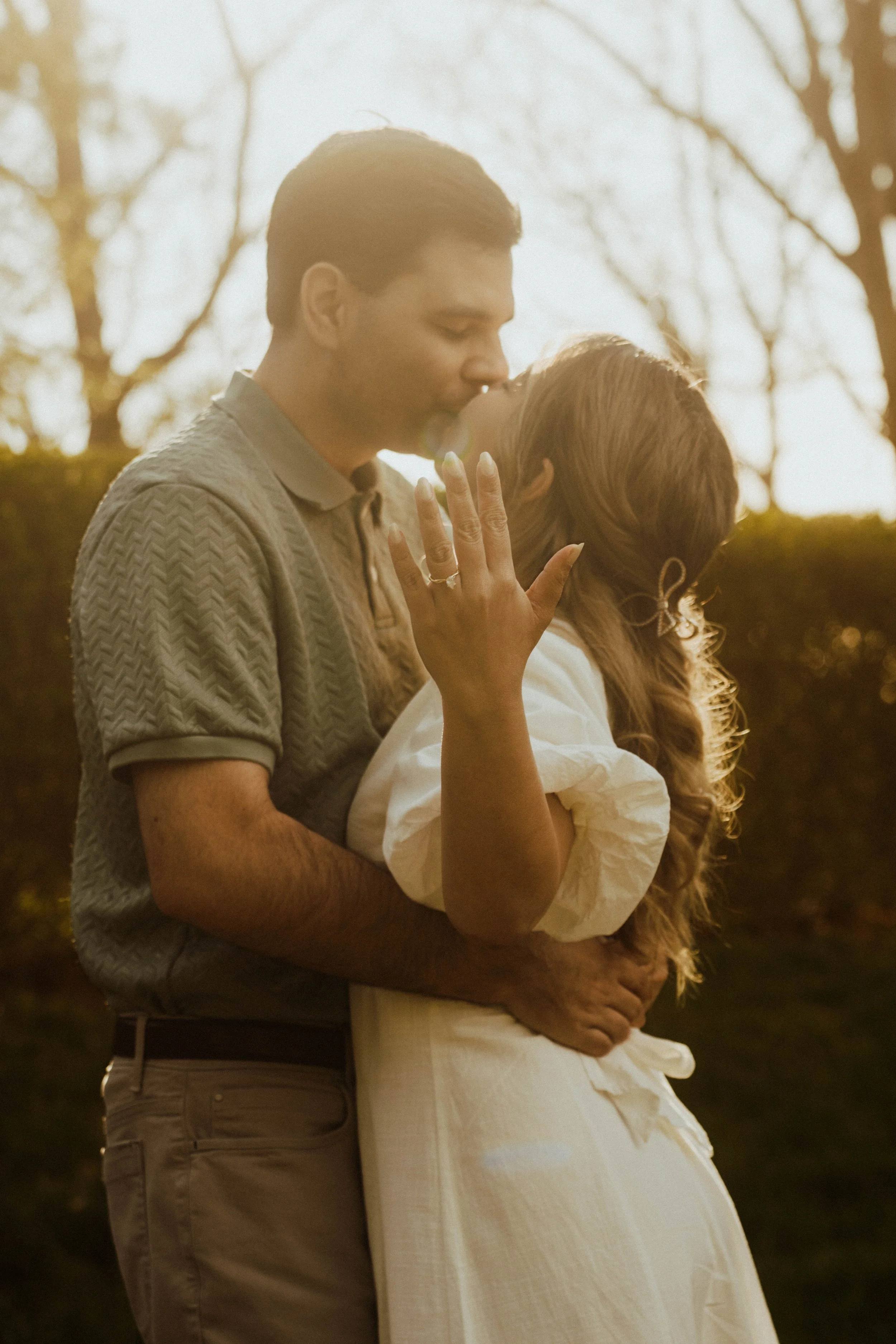 A couple standing outdoors embracing each other, with the man gently kissing the woman's forehead during sunset.