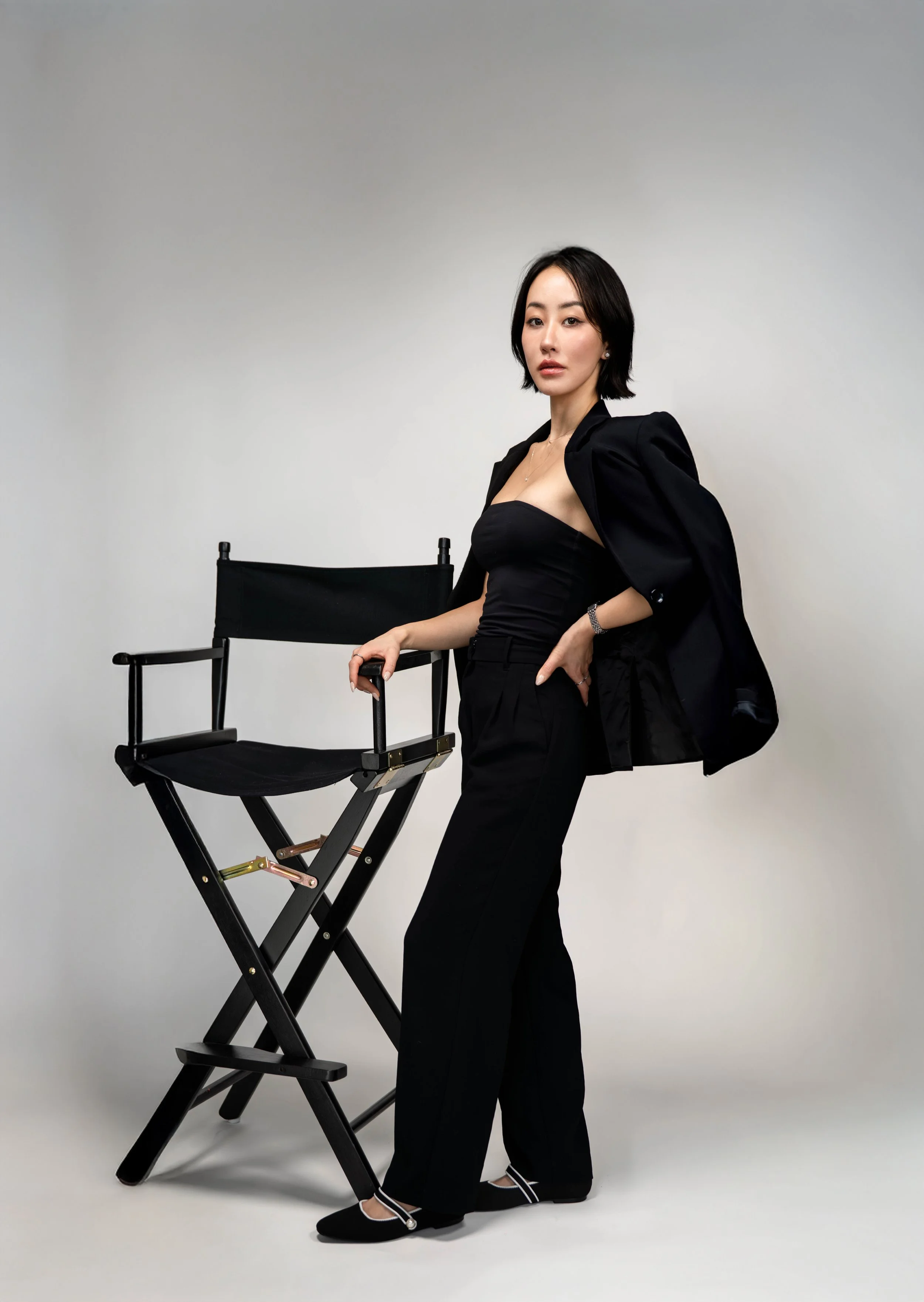 A woman in black pants and a strapless top, with a black blazer draped over her shoulders, standing next to a black director's chair against a plain white background.