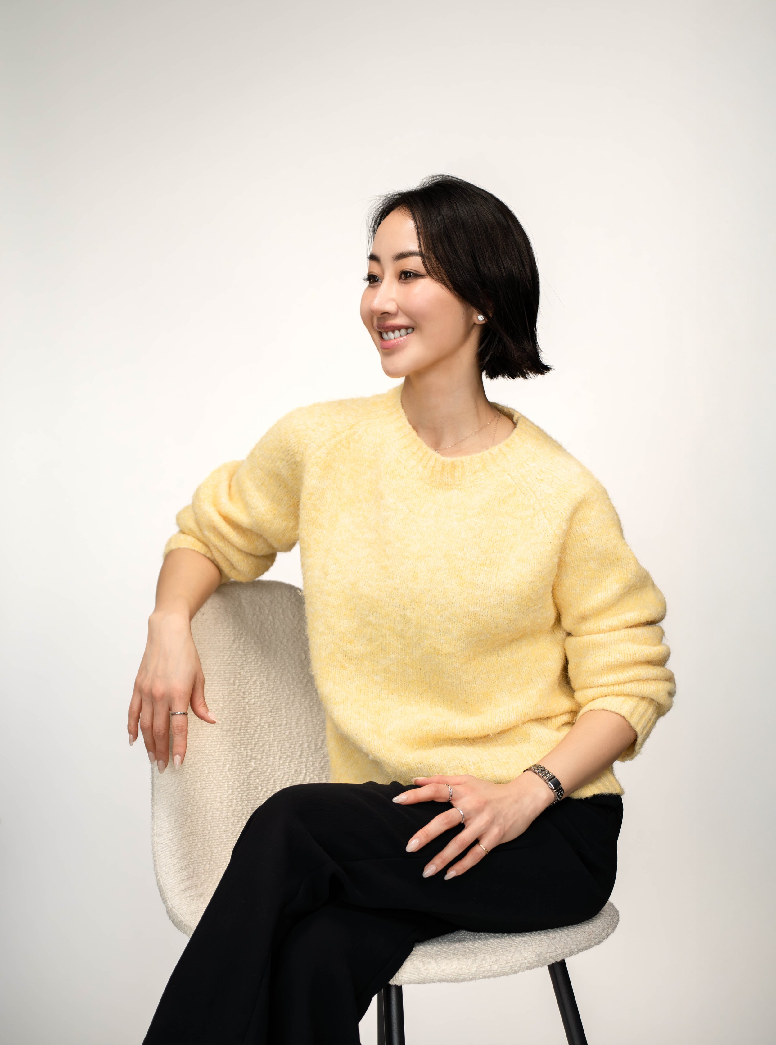 A woman sitting on a beige chair, wearing a yellow sweater and black pants, smiling and looking to the side against a plain white background.