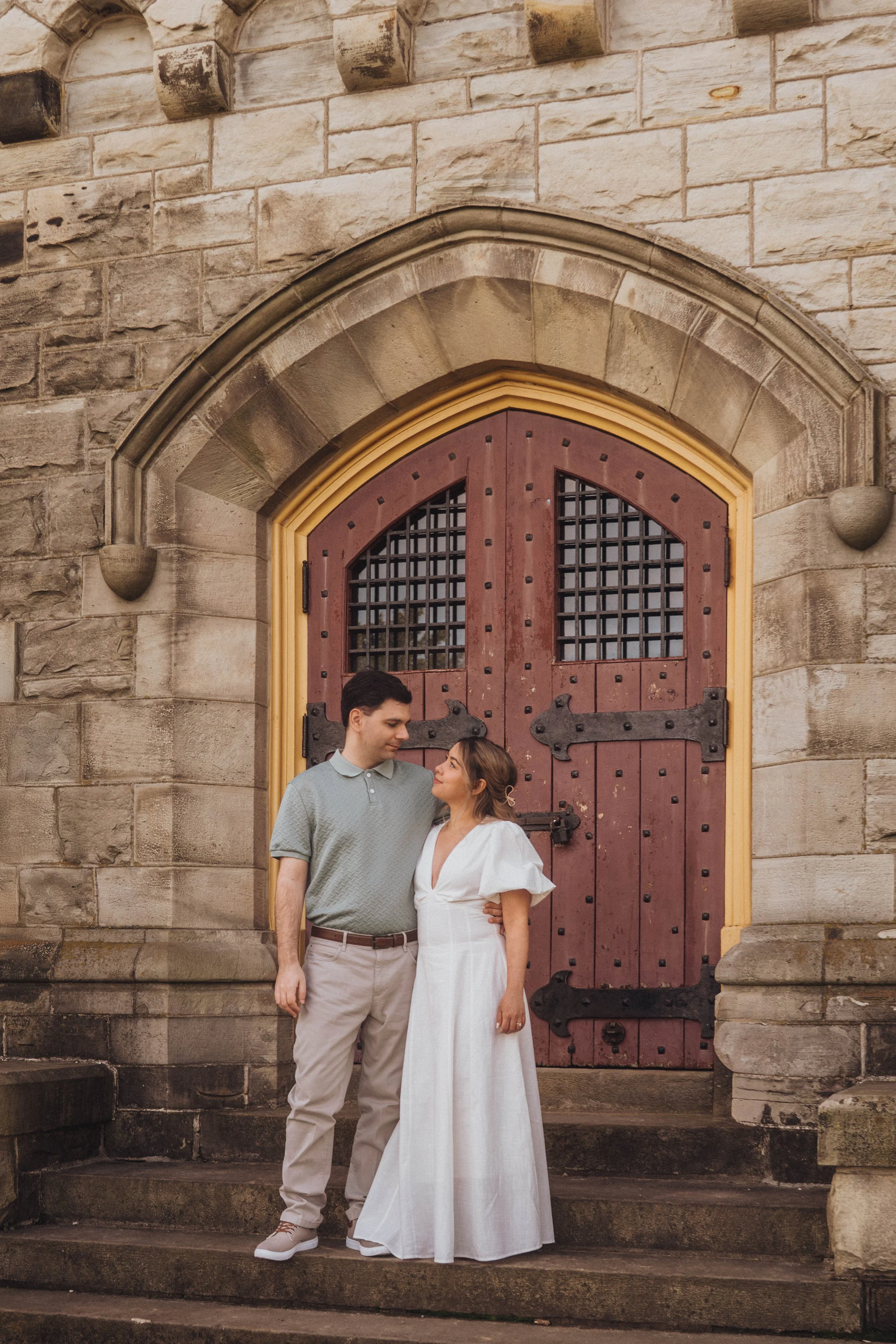 A couple standing close together in front of a wooden door with a rounded arch, attached to a stone building. The man is wearing a light gray polo shirt and beige pants, and the woman is in a white dress. They are looking at each other with affection