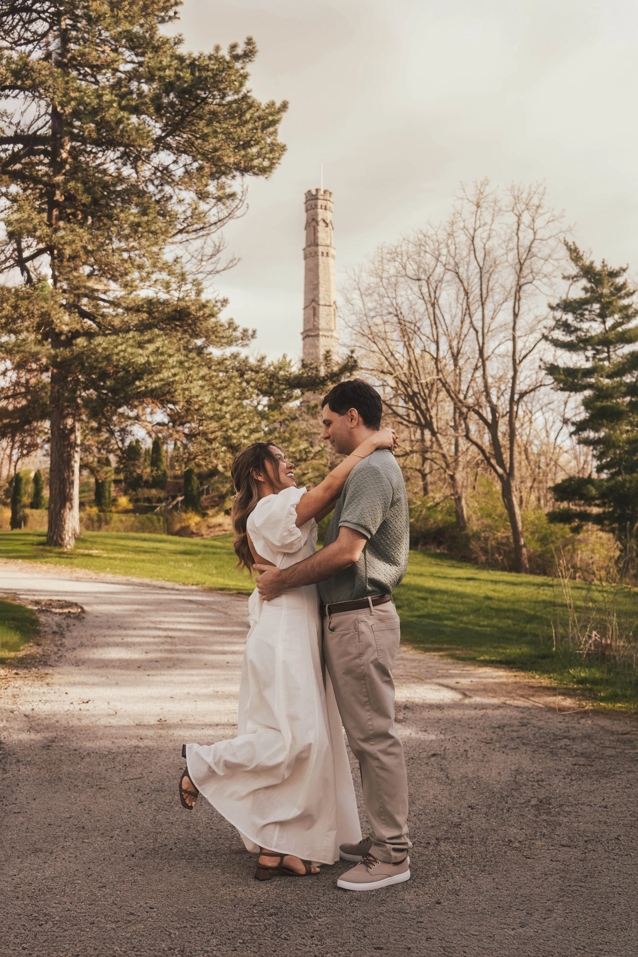A couple dancing in an outdoor park surrounded by trees with a tall tower in the background, during the daytime.