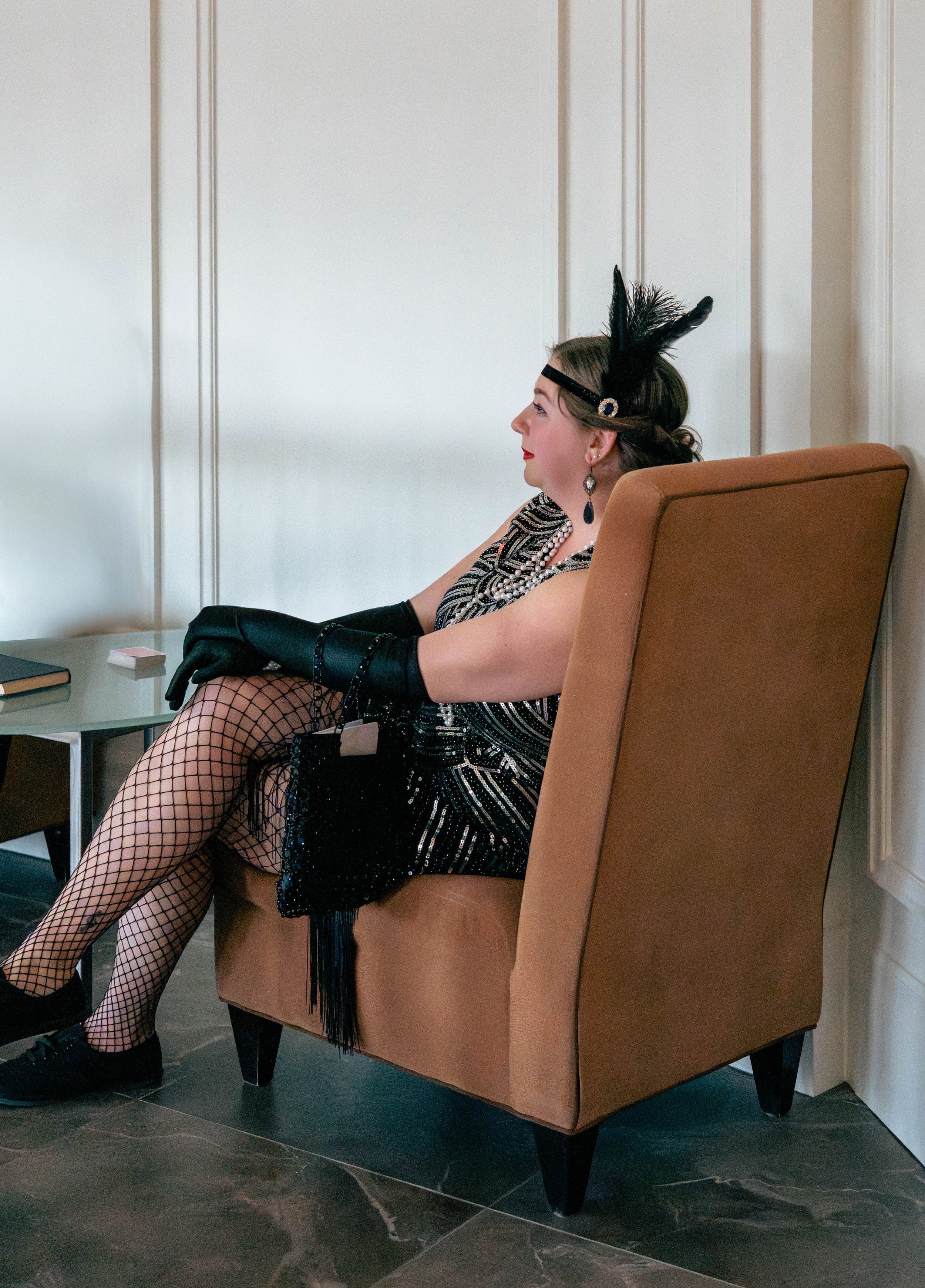 A woman dressed in 1920s flapper costume, sitting on a tan armchair. She's wearing a black sequin dress, fishnet stockings, black gloves, a headband with feathers, and jewelry, with a side profile view.