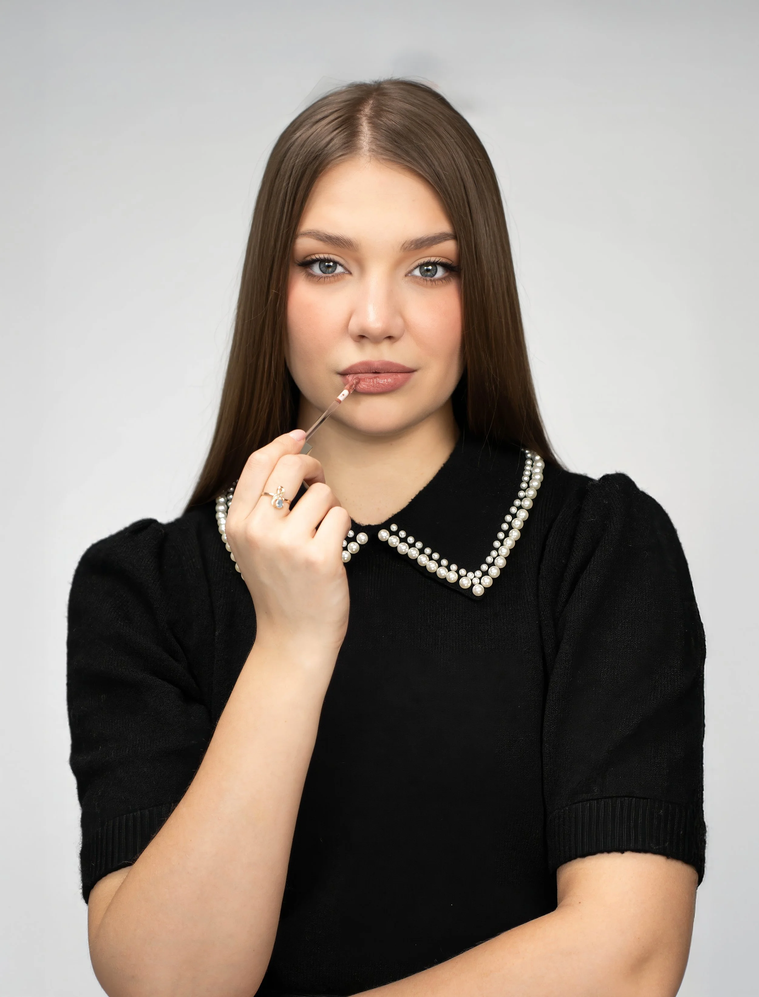A woman with long brown hair and blue eyes is holding a makeup brush near her lips, wearing a black top with pearl details on the collar.