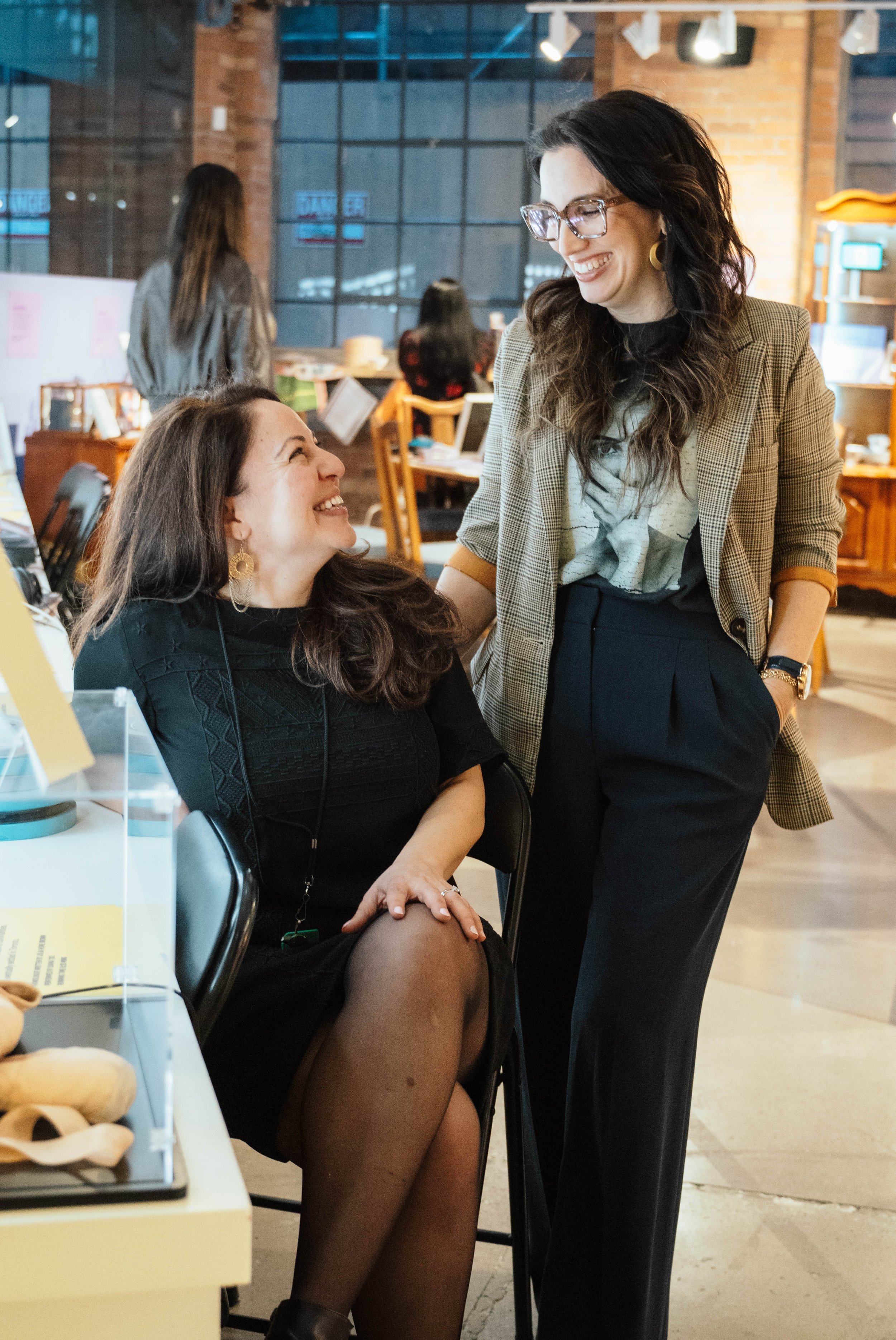 Two women are smiling and talking in an indoor setting, with one sitting on a chair and the other standing. The woman sitting is wearing a black dress and sheer black tights, and the woman standing is dressed in a plaid blazer and dark pants.