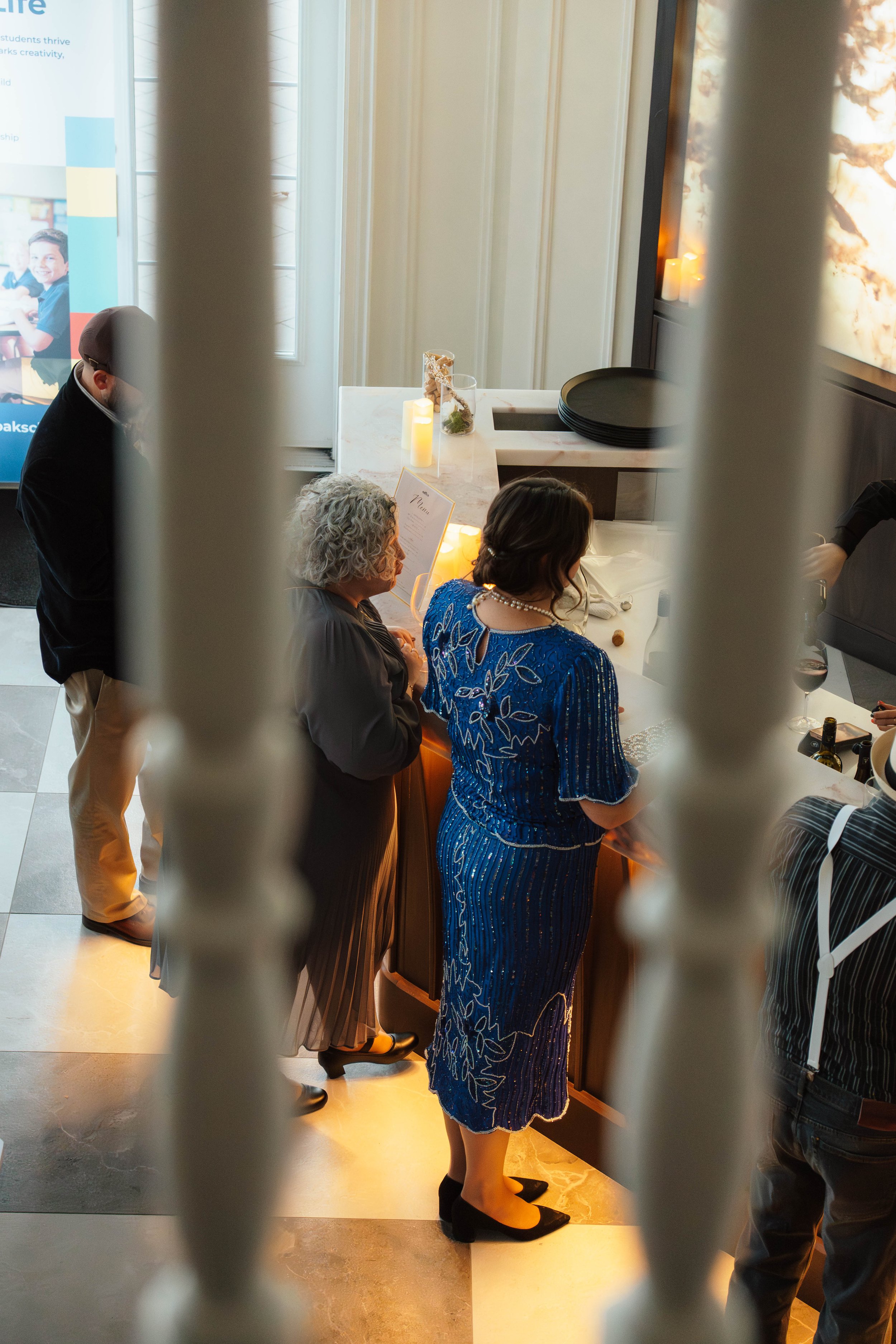 People standing behind a white kitchen counter, viewed through a railing, possibly at a restaurant or event. The scene is lit with warm lighting.