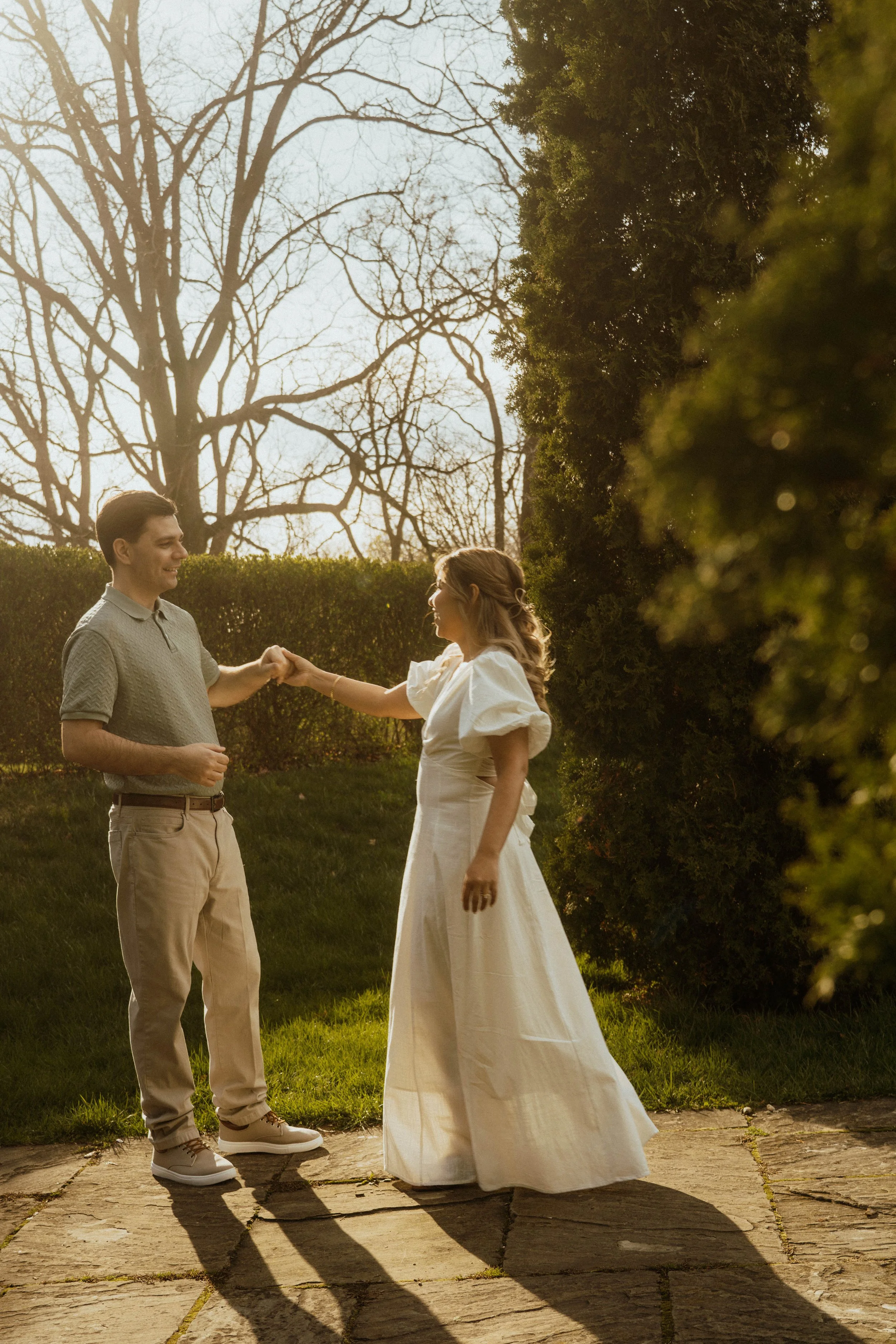 A man and woman holding hands outdoors on a sunny day, with trees and bushes in the background.