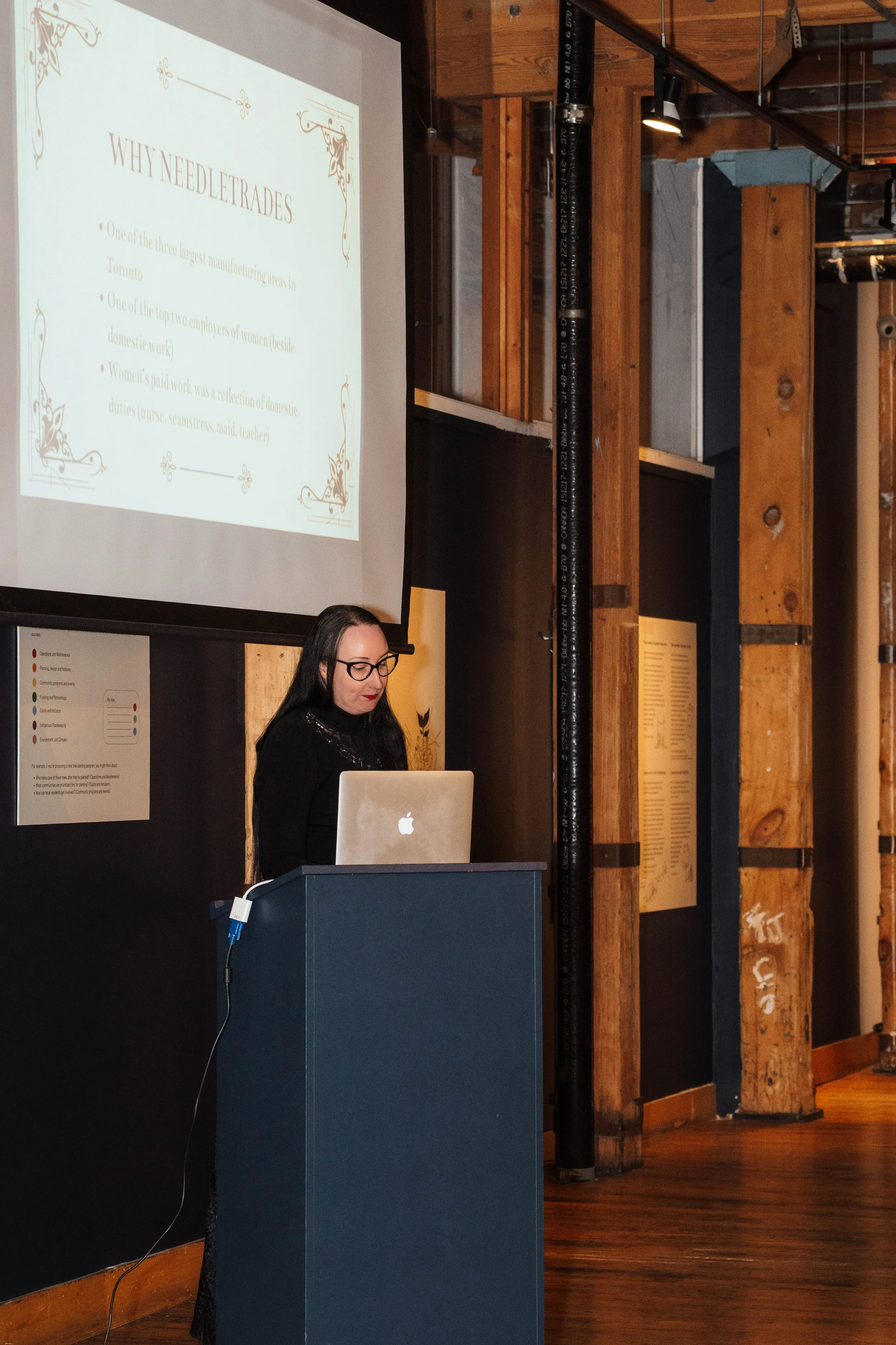 Woman giving a presentation at a podium with a laptop, with a large screen displaying a slide titled "WHY NEEDLETRADES" in a room with wooden beams.